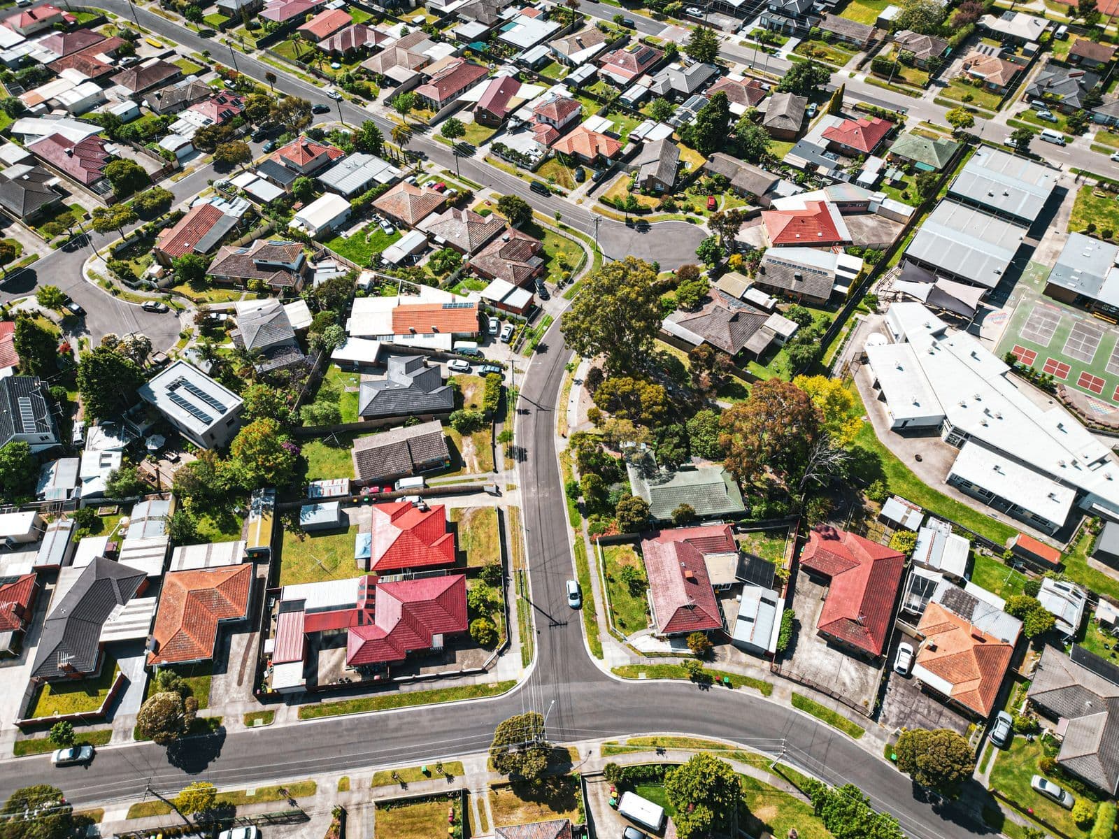 An aerial view of an Australian suburb, with tiled-roof houses, tree-lined streets and cars parked along the kerbs.