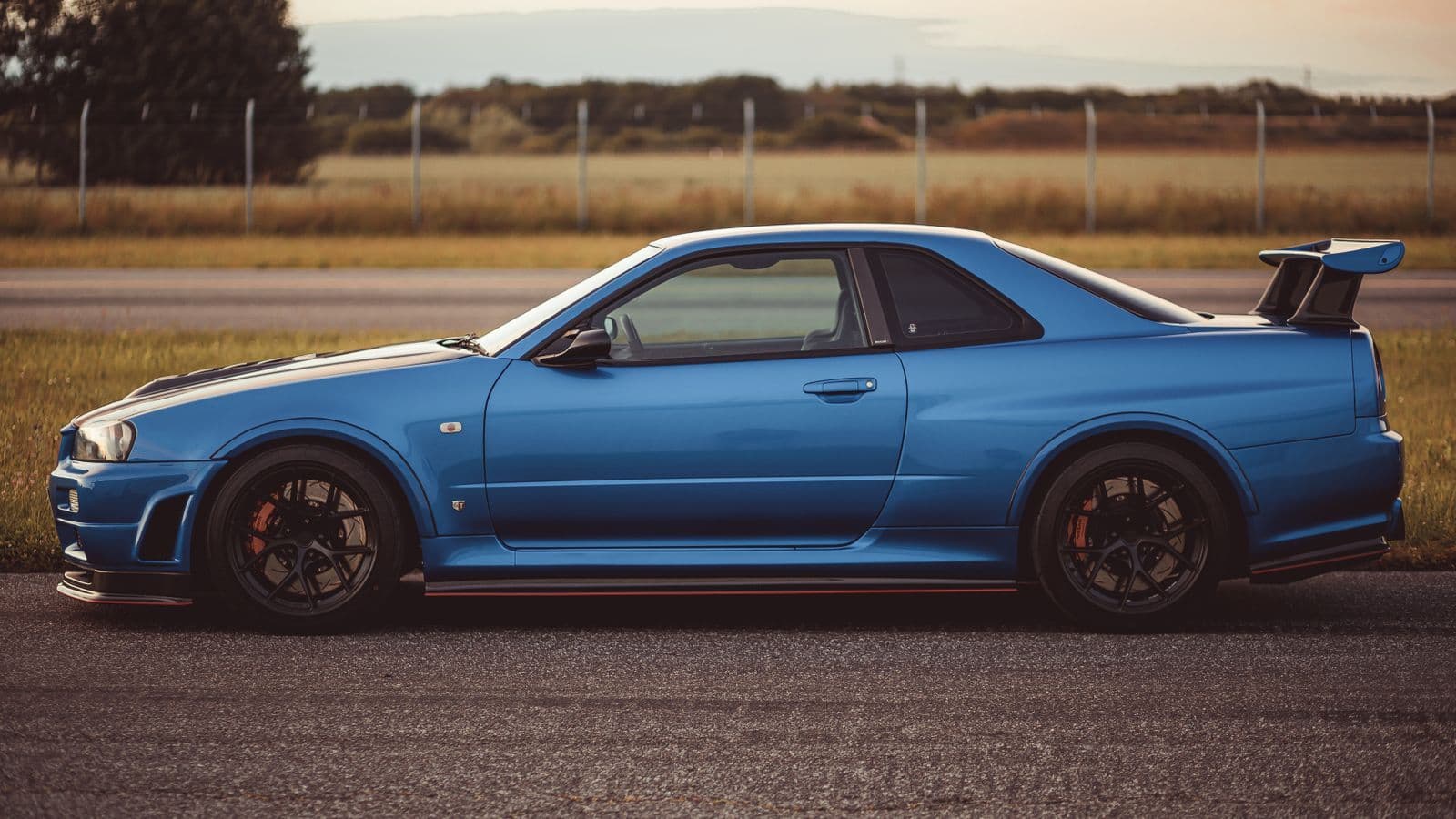 Side view of a blue Nissan Skyline GT-R R34 with a rear wing, parked on a track apron at dusk.