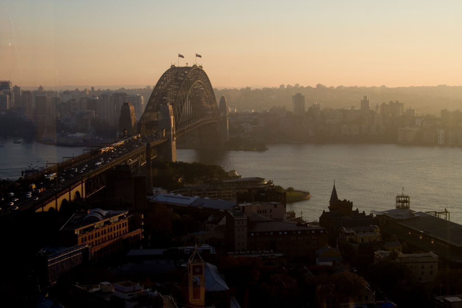 Sydney Harbour Bridge at sunset seen from above, with traffic queued along the approach road and the harbour and city beyond.