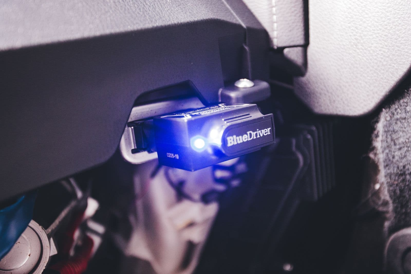 A diagnostic scan tool with a glowing blue light plugged into the OBD port beneath a car's dashboard.