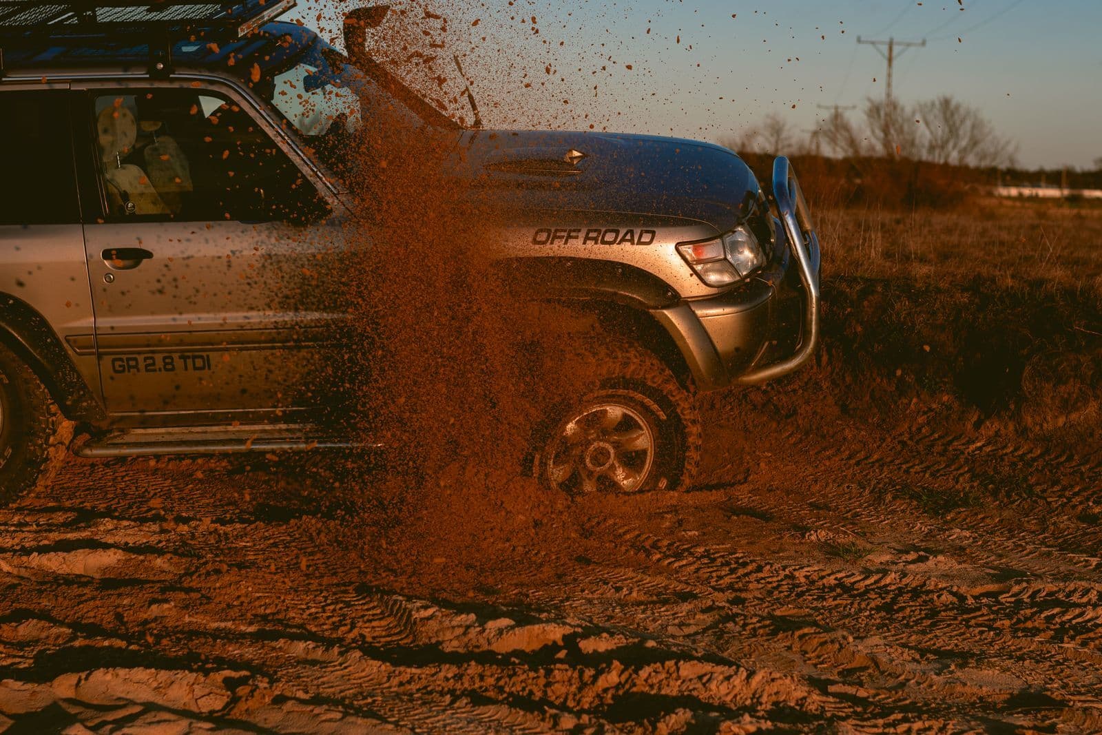 A silver four-wheel drive throws up a spray of red dirt as it drives through a muddy track at sunset.