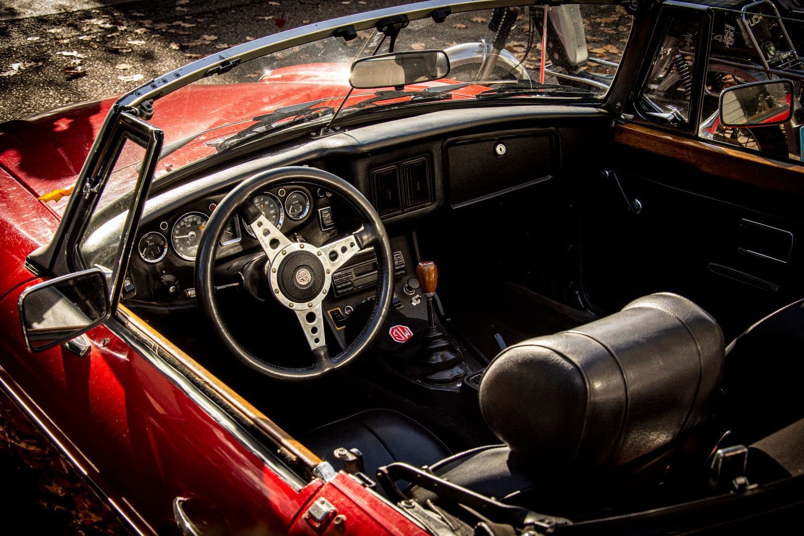 The cockpit of a red classic MG convertible, with a wood-rimmed alloy steering wheel and round analogue gauges, lit by low sunlight.