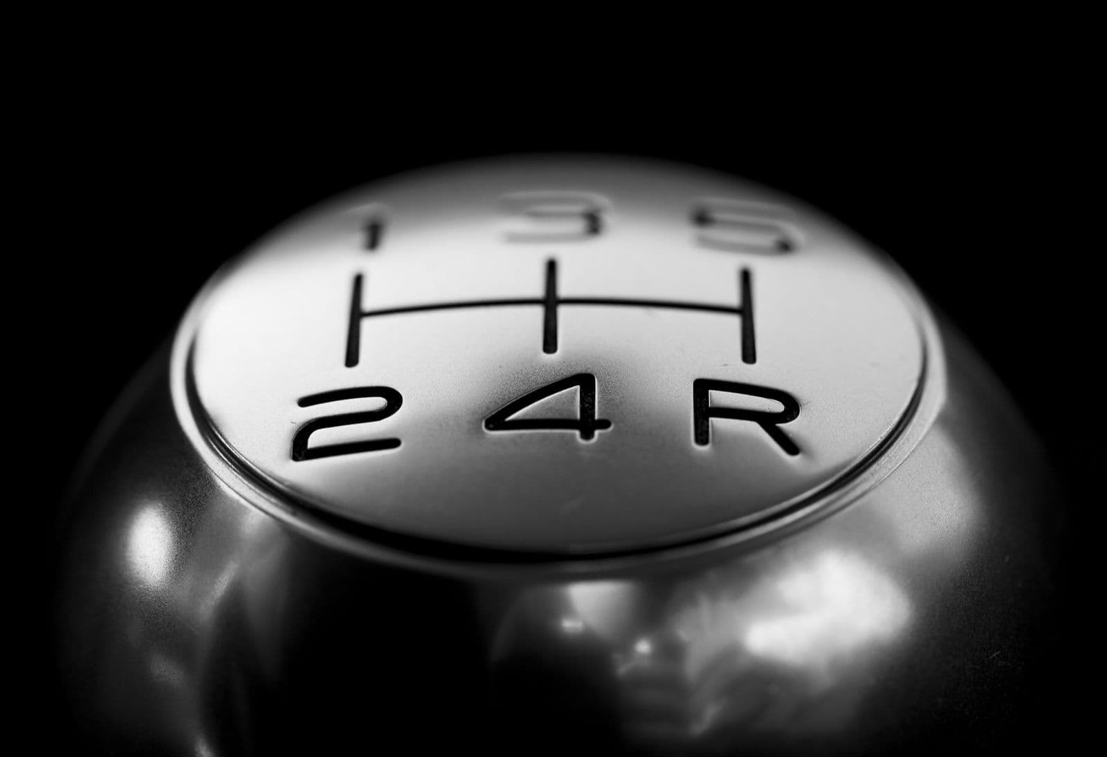 A black-and-white close-up of a polished metal gear knob engraved with a five-speed manual shift pattern.