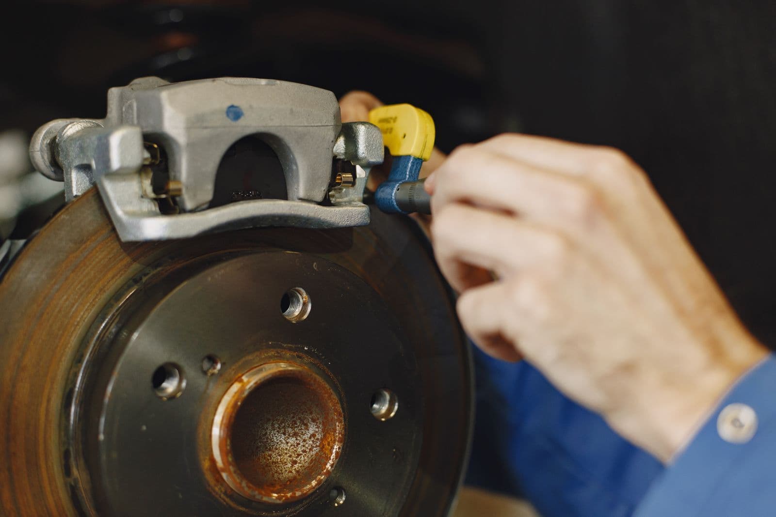 A mechanic's hand works on a brake caliper mounted over a rust-flecked brake disc with the wheel removed.