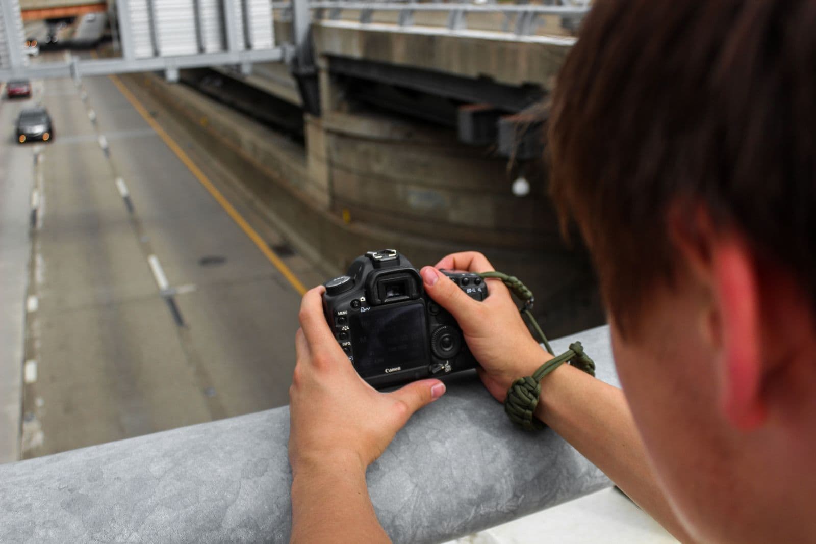 A person holds a DSLR camera over a concrete railing above a multi-lane road, with cars passing below.