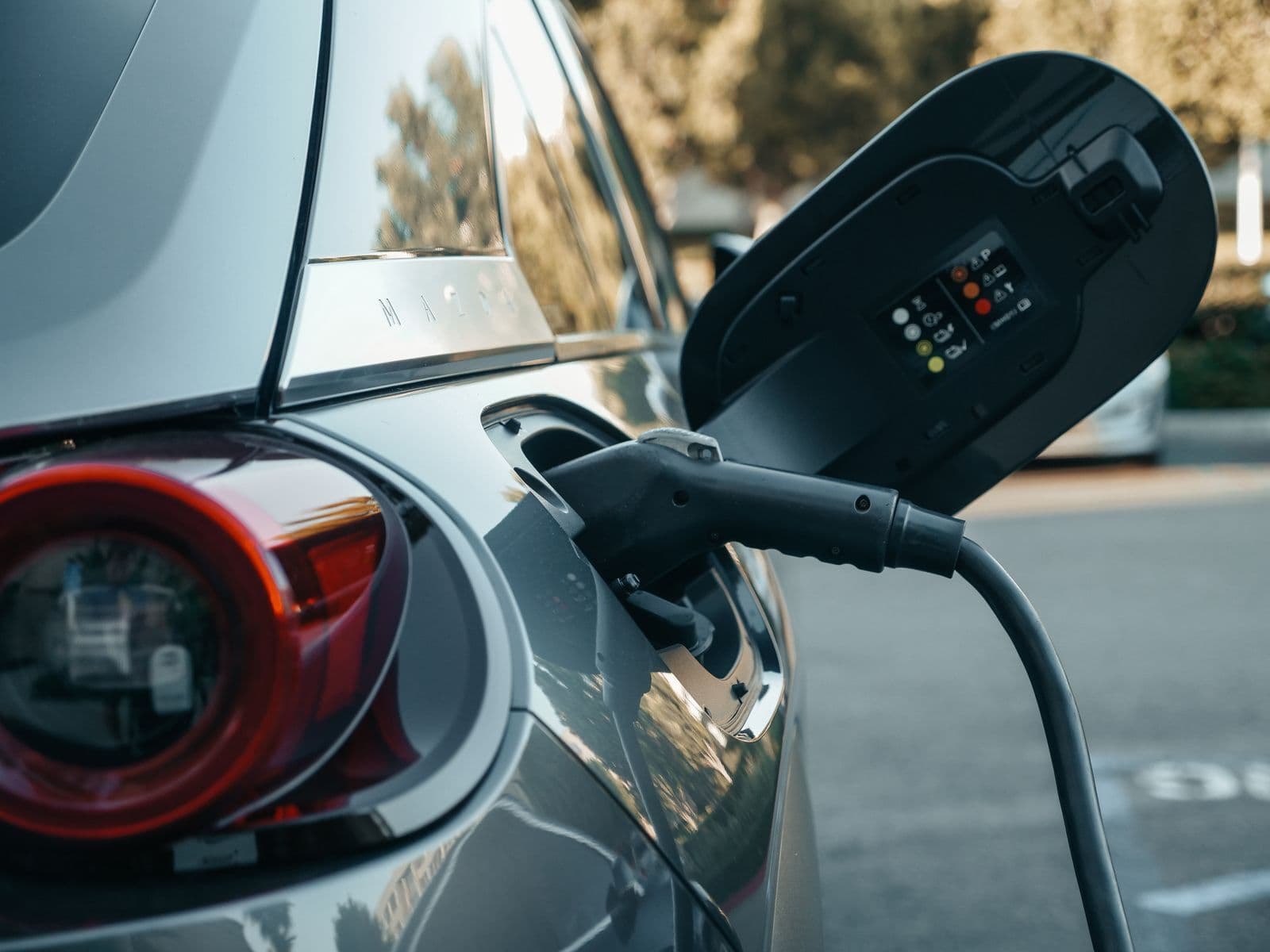 A charging cable plugged into the open charge port of a silver electric car, with coloured status lights glowing on the port flap.