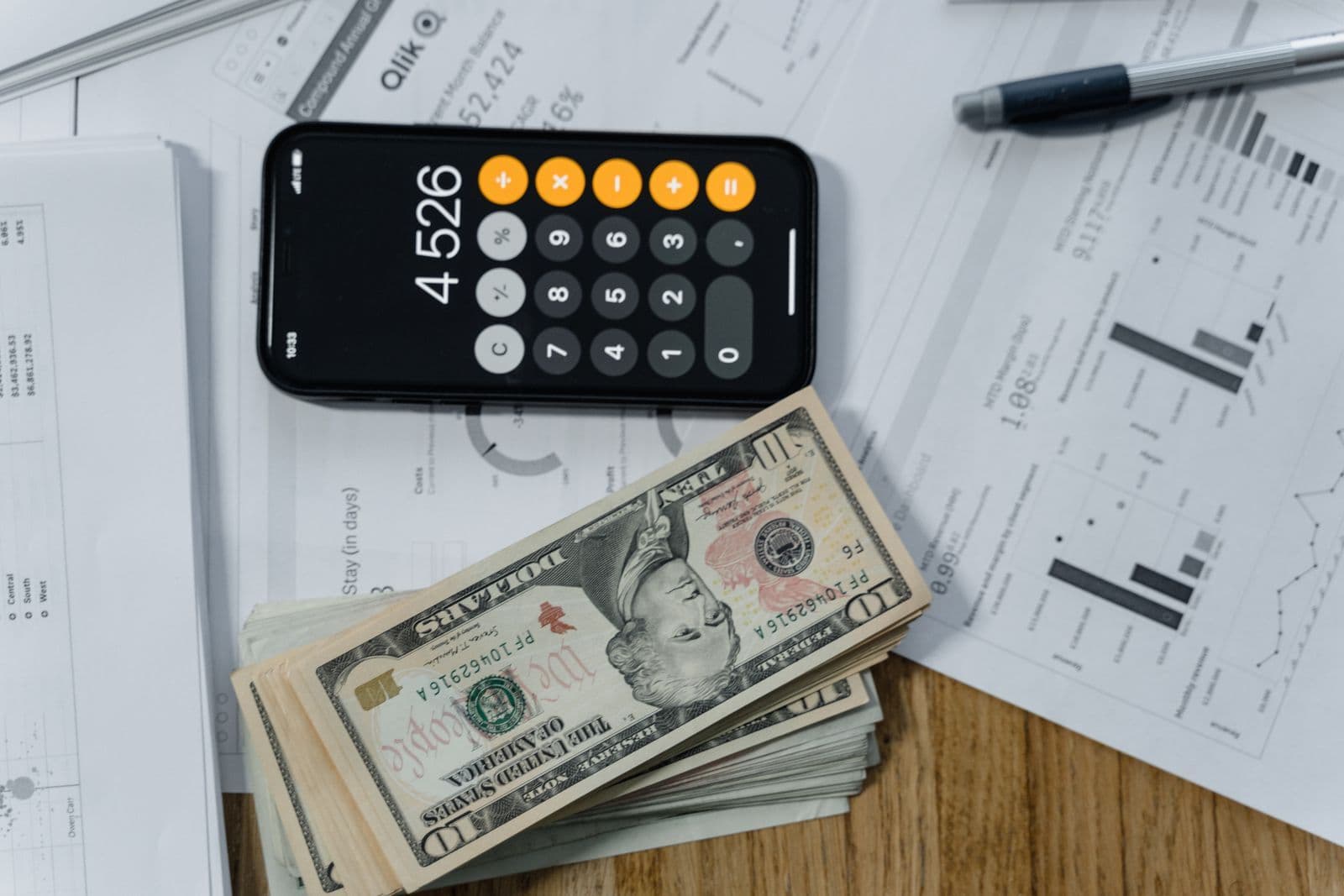 A phone showing a calculator app beside a thick stack of banknotes and printed charts on a desk.