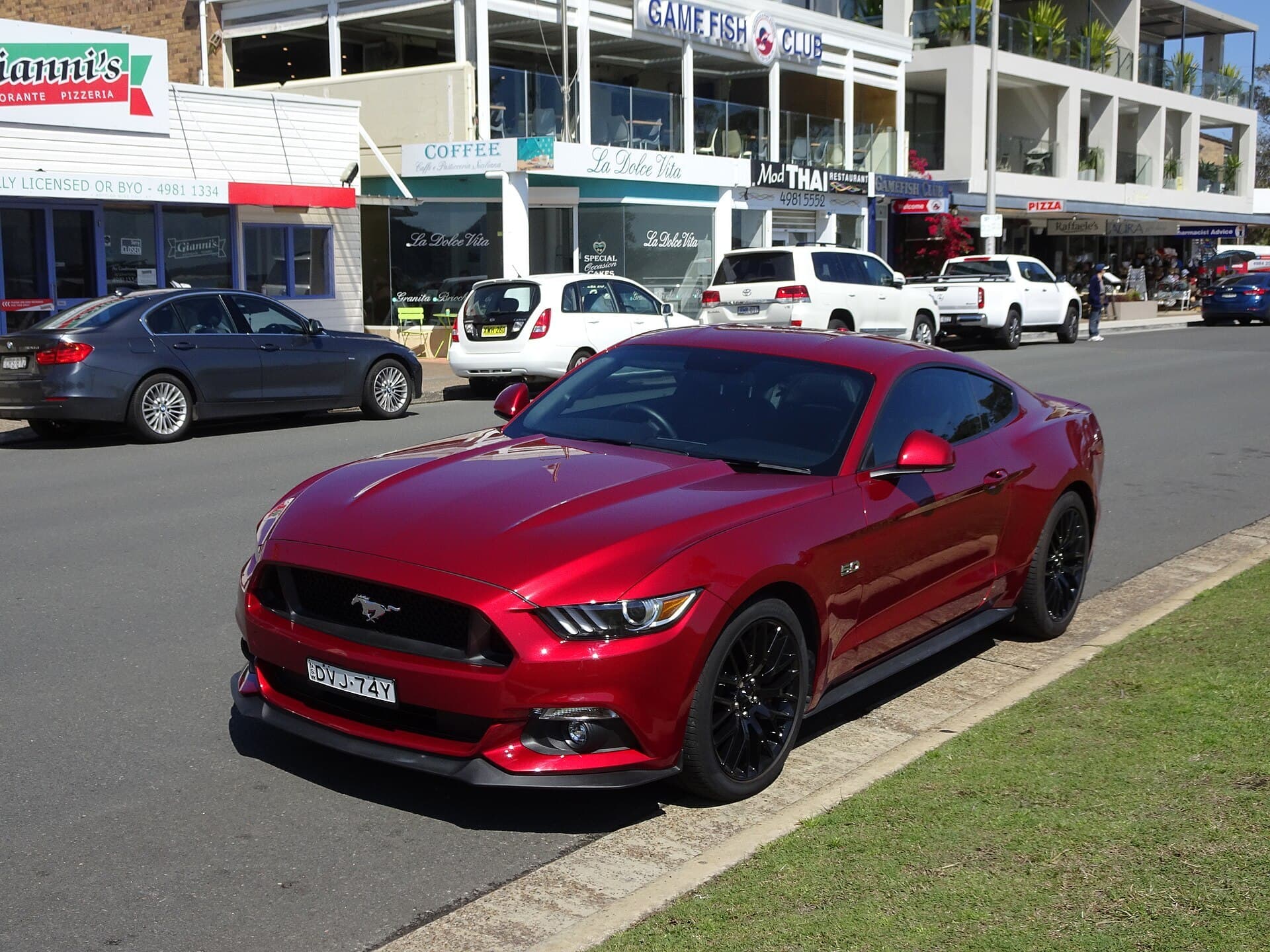 A red Ford Mustang GT coupe with black wheels parked at the kerb of a beachside shopping street with cafes and restaurants behind it.