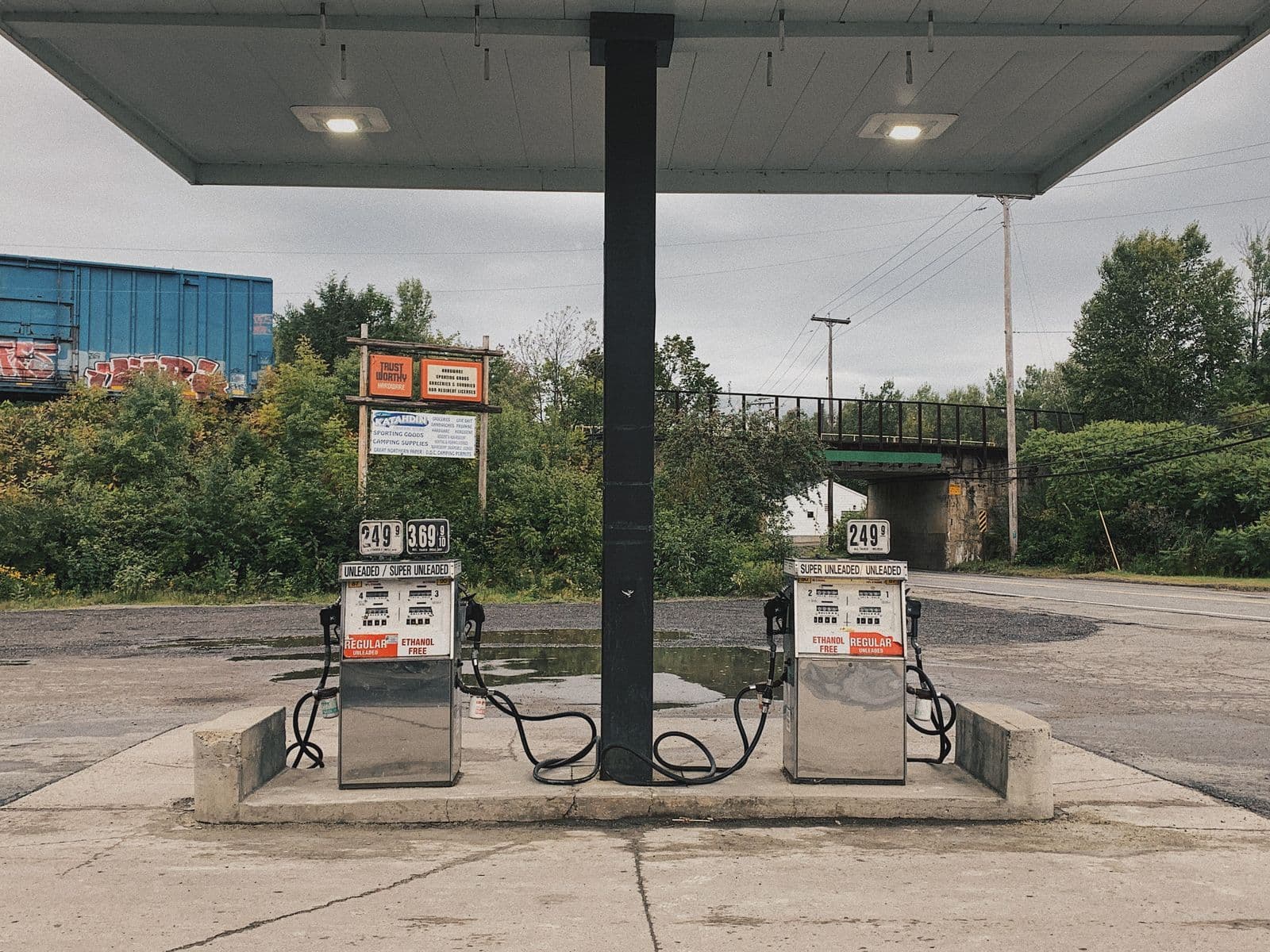 Two fuel pumps with their prices displayed, standing under the canopy of a small roadside service station on an overcast day.