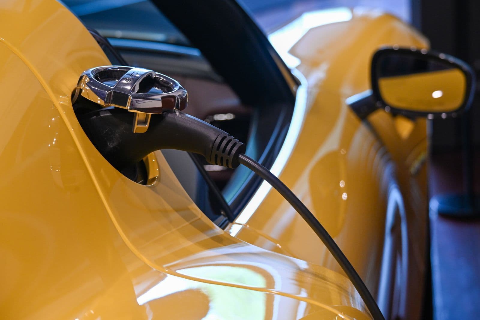 A black charging connector plugged into the charge port on the front wing of a glossy yellow car, with its wing mirror behind.