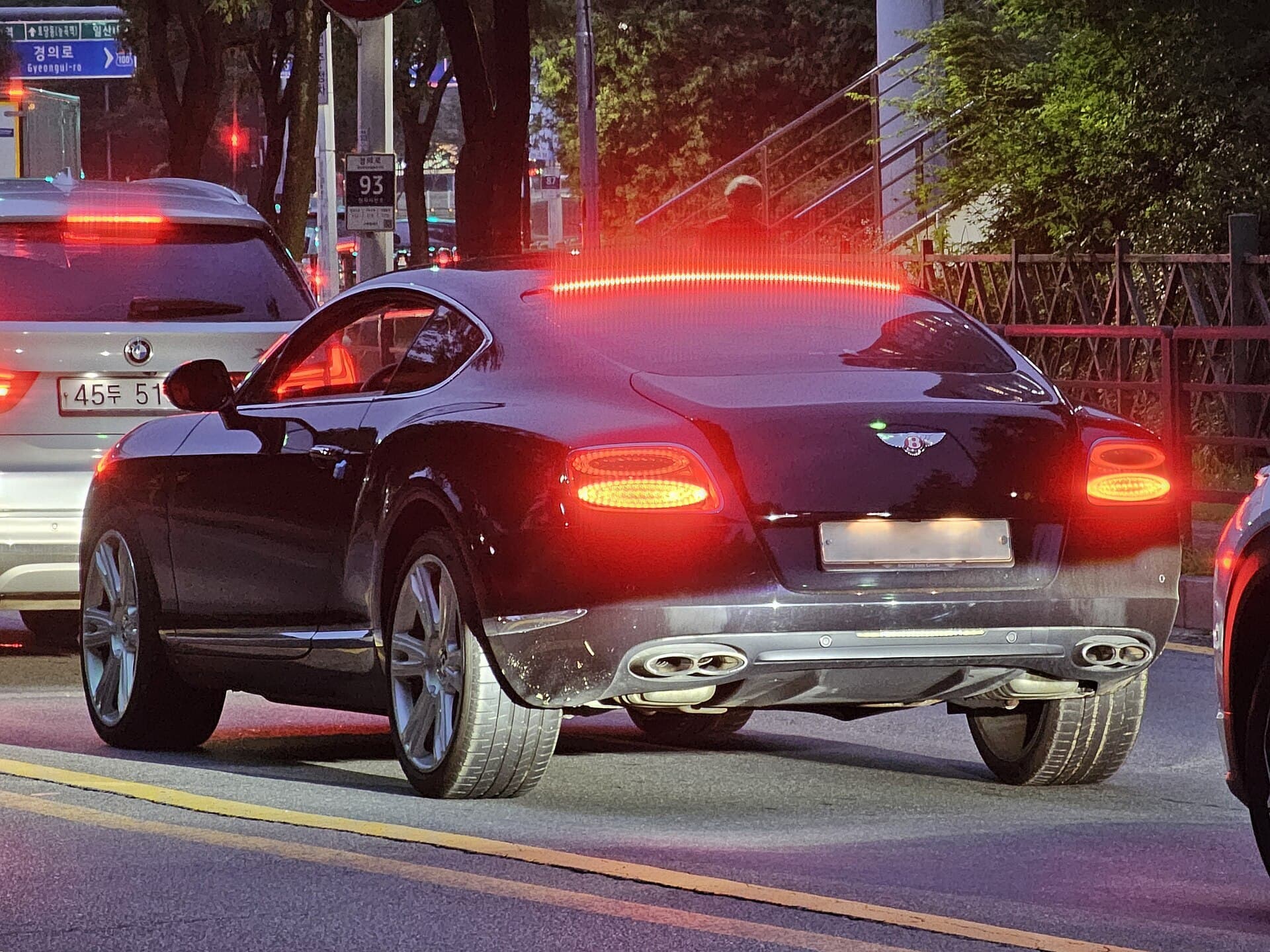 A dark Bentley Continental GT coupe seen from behind on a city street at dusk, its brake lights on.