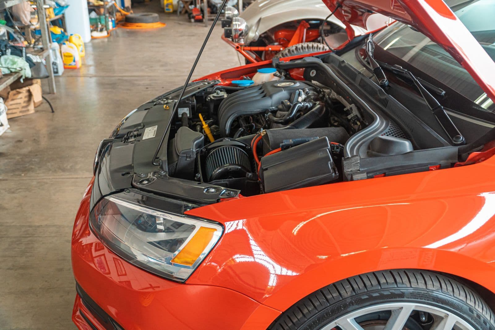 The open bonnet of an orange car in a workshop, showing an engine bay crowded with black plastic ducts, covers and a translucent coolant reservoir.