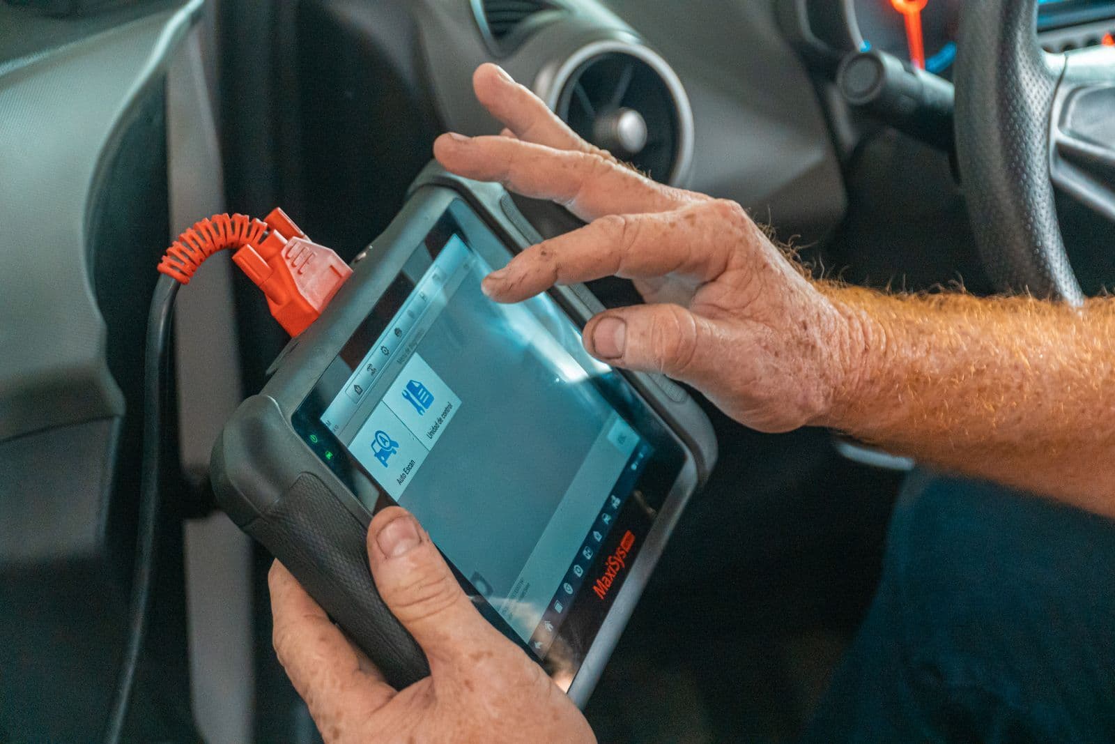 A technician holds a handheld diagnostic tablet connected by an orange cable to a car's dashboard socket.