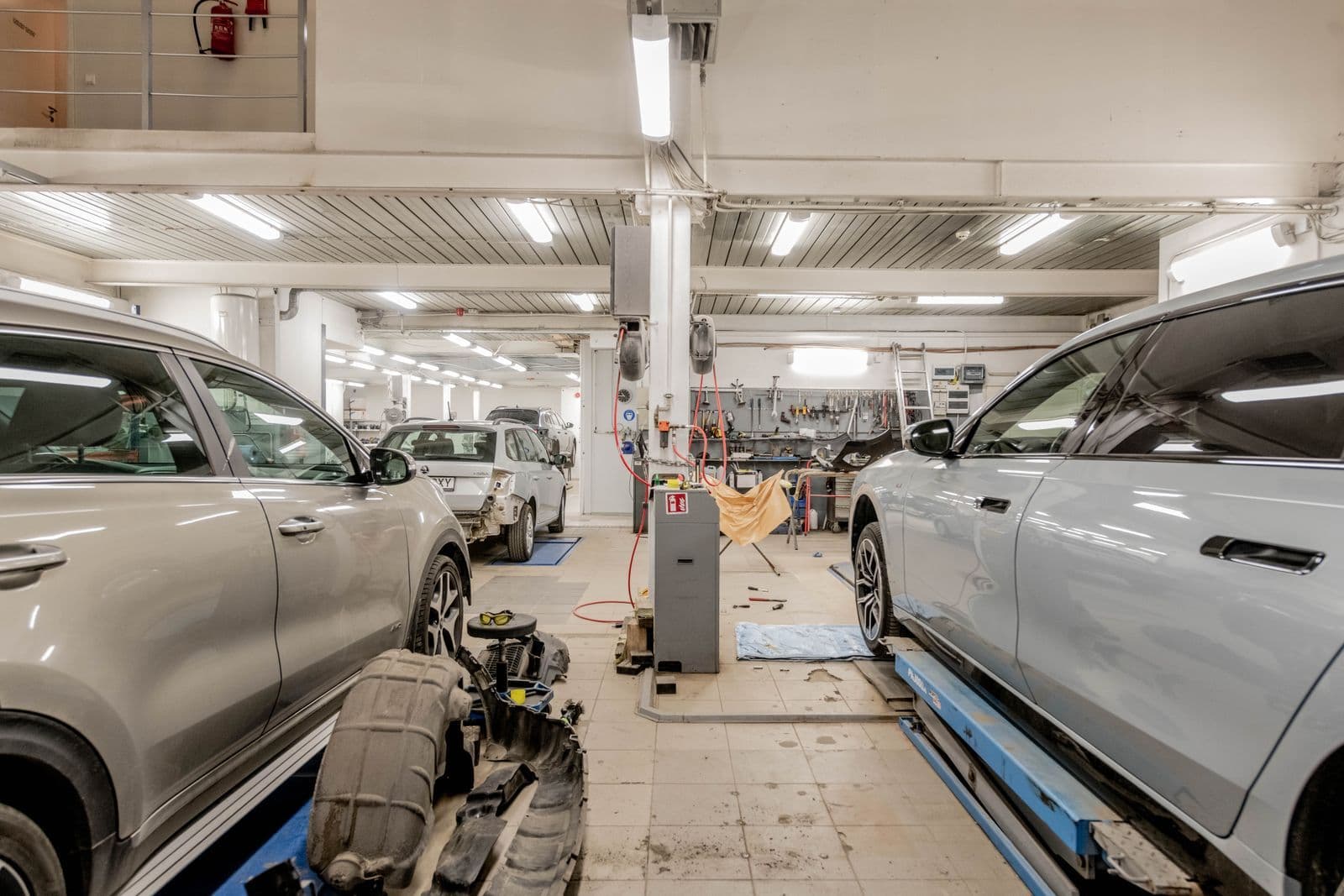 The inside of a busy service workshop, with cars raised on hoists either side of a strip-lit central aisle and tools racked along the back wall.