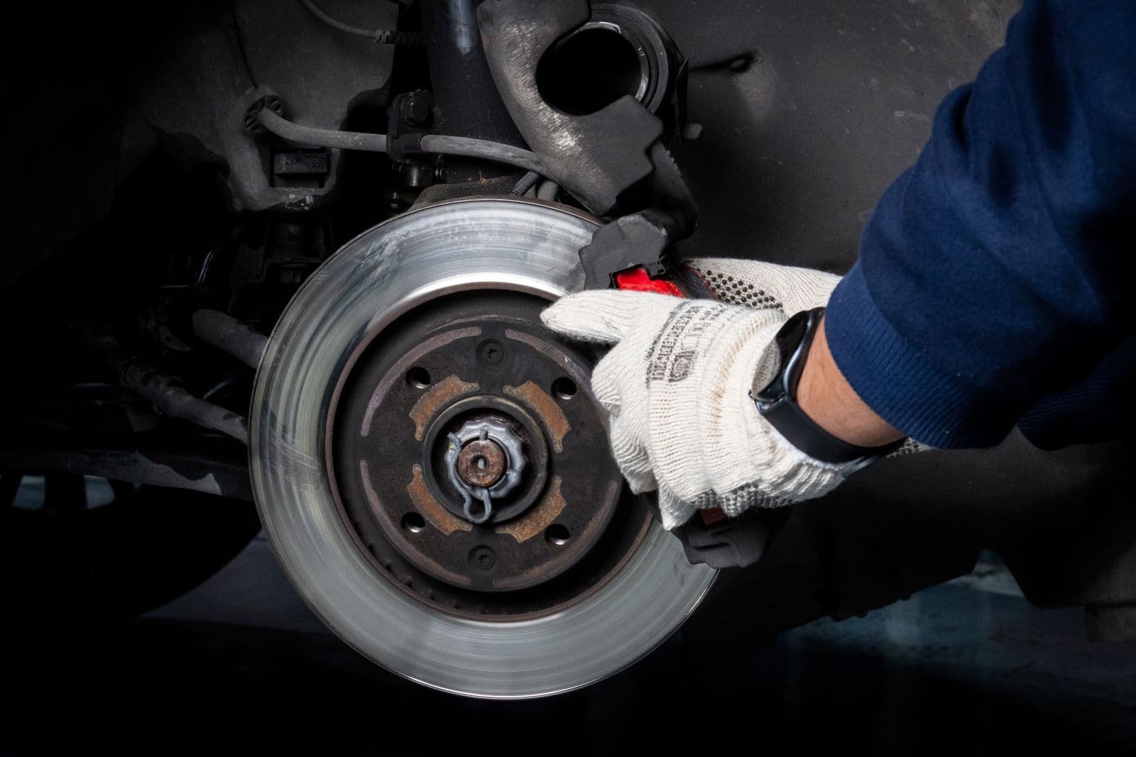 A gloved hand resting on a worn brake disc and caliper under a car with the wheel removed.