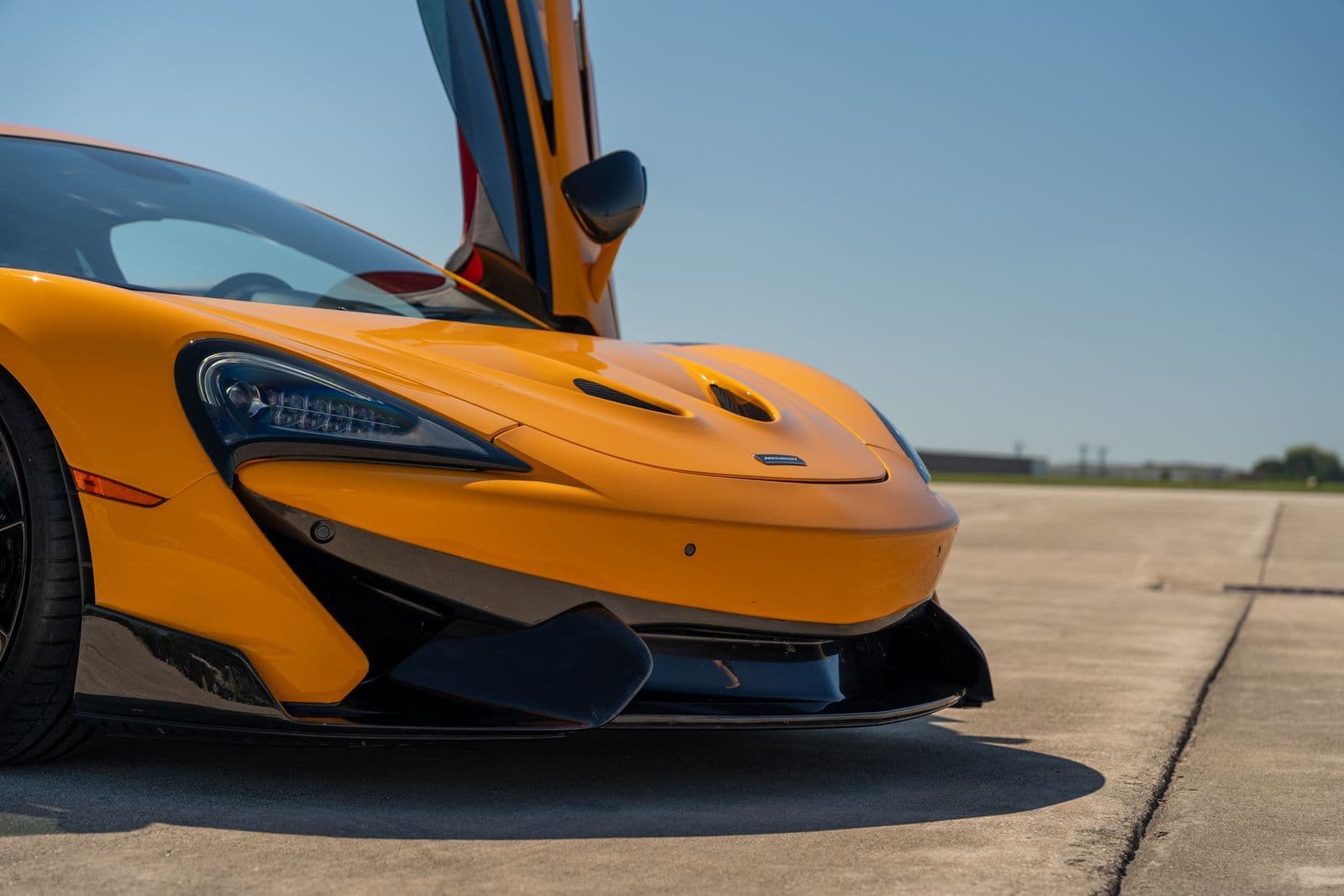 Close-up of the front of an orange McLaren sports car with its dihedral door raised, parked on an airfield apron.