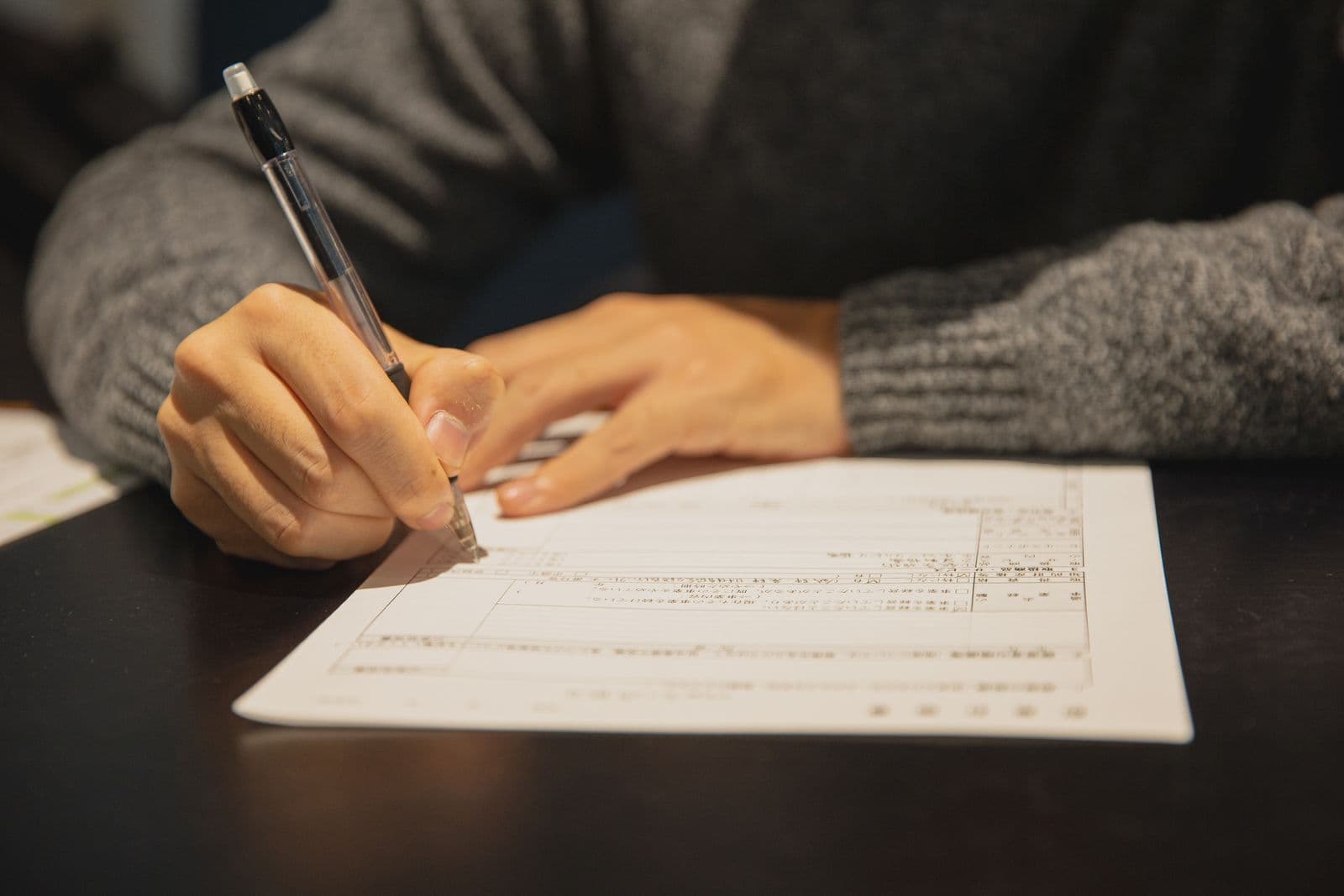 Close-up of a person in a grey jumper filling in a printed form with a pen at a dark table.