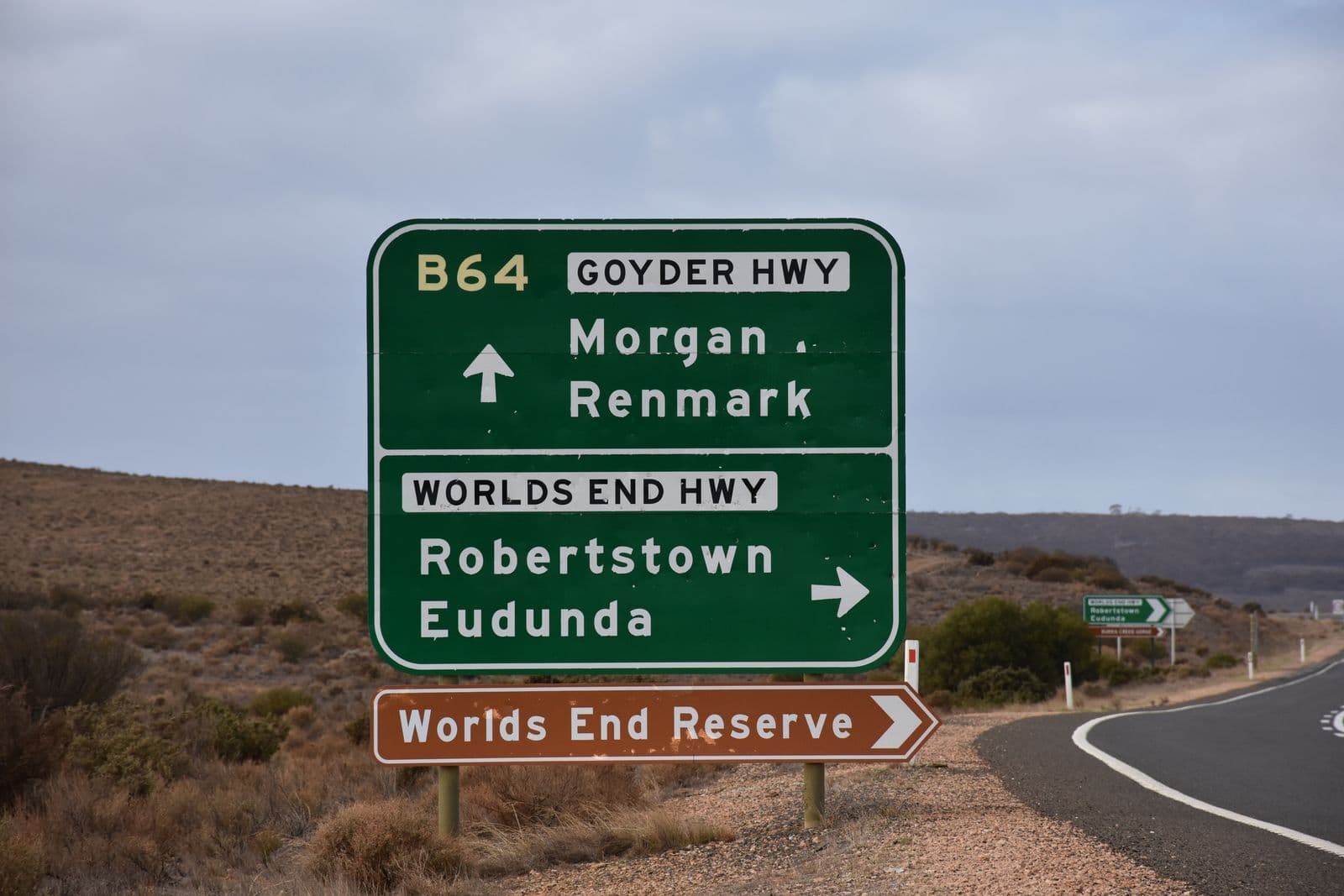 A green Australian highway direction sign beside a sealed rural road, pointing to Morgan, Renmark, Robertstown and Eudunda across dry scrub.