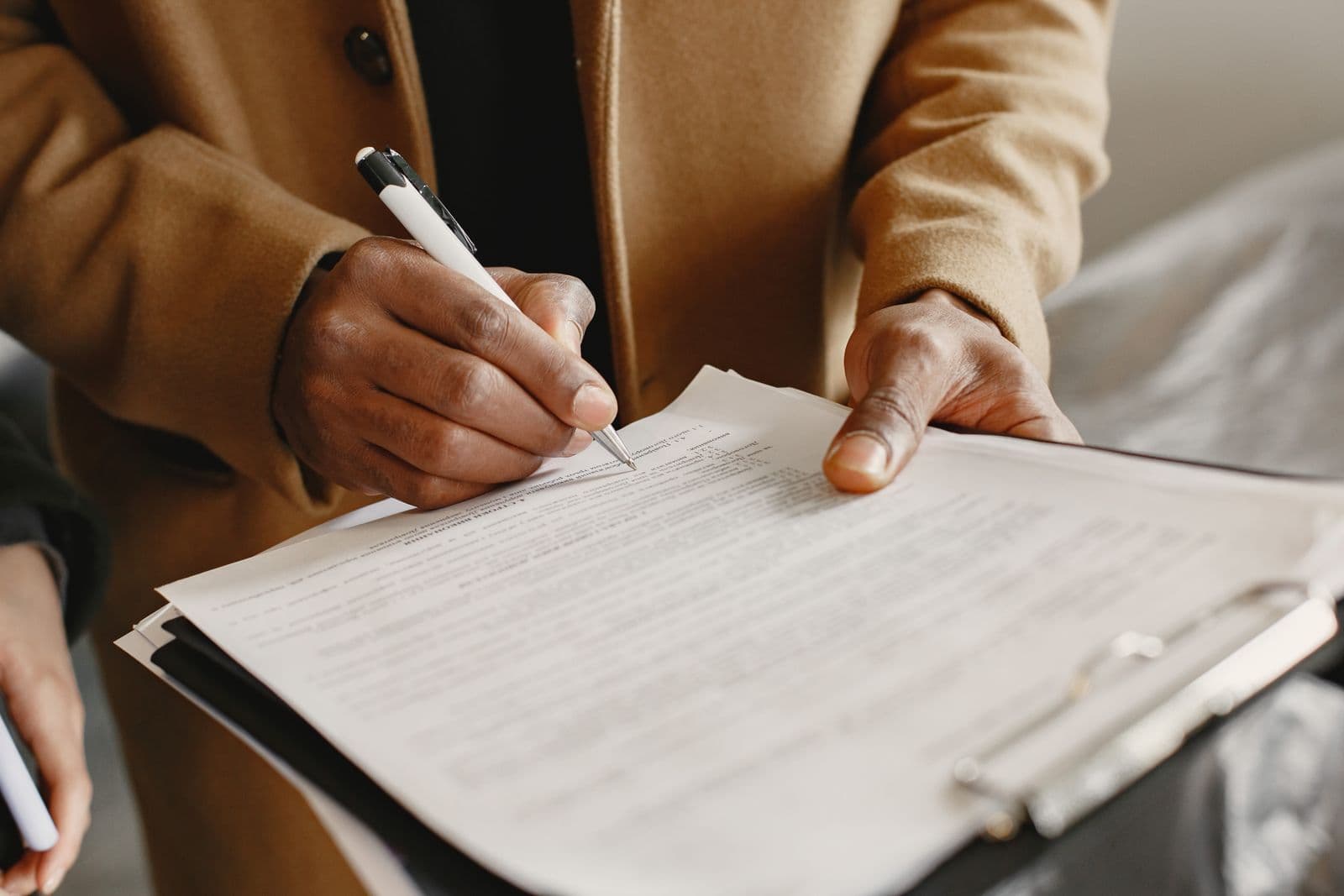 A person in a tan coat signing a printed agreement held on a clipboard, with the pen mid-stroke on the signature line.