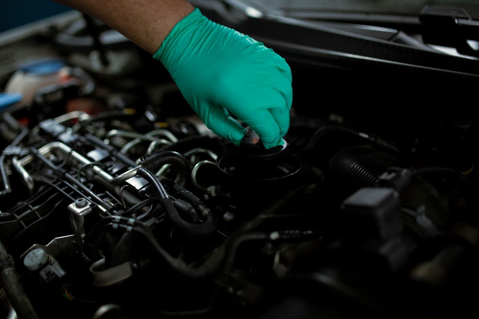 A hand in a green nitrile glove opening the oil filler cap on top of a car engine.