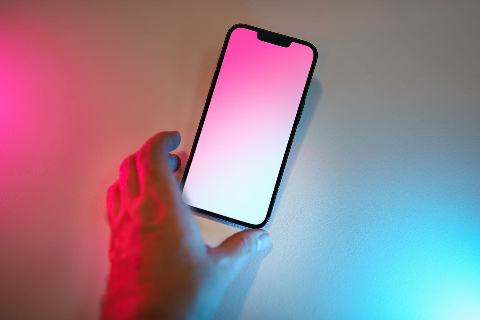 A hand reaches for a smartphone with a glowing blank screen, lit by pink and blue light against a pale wall.