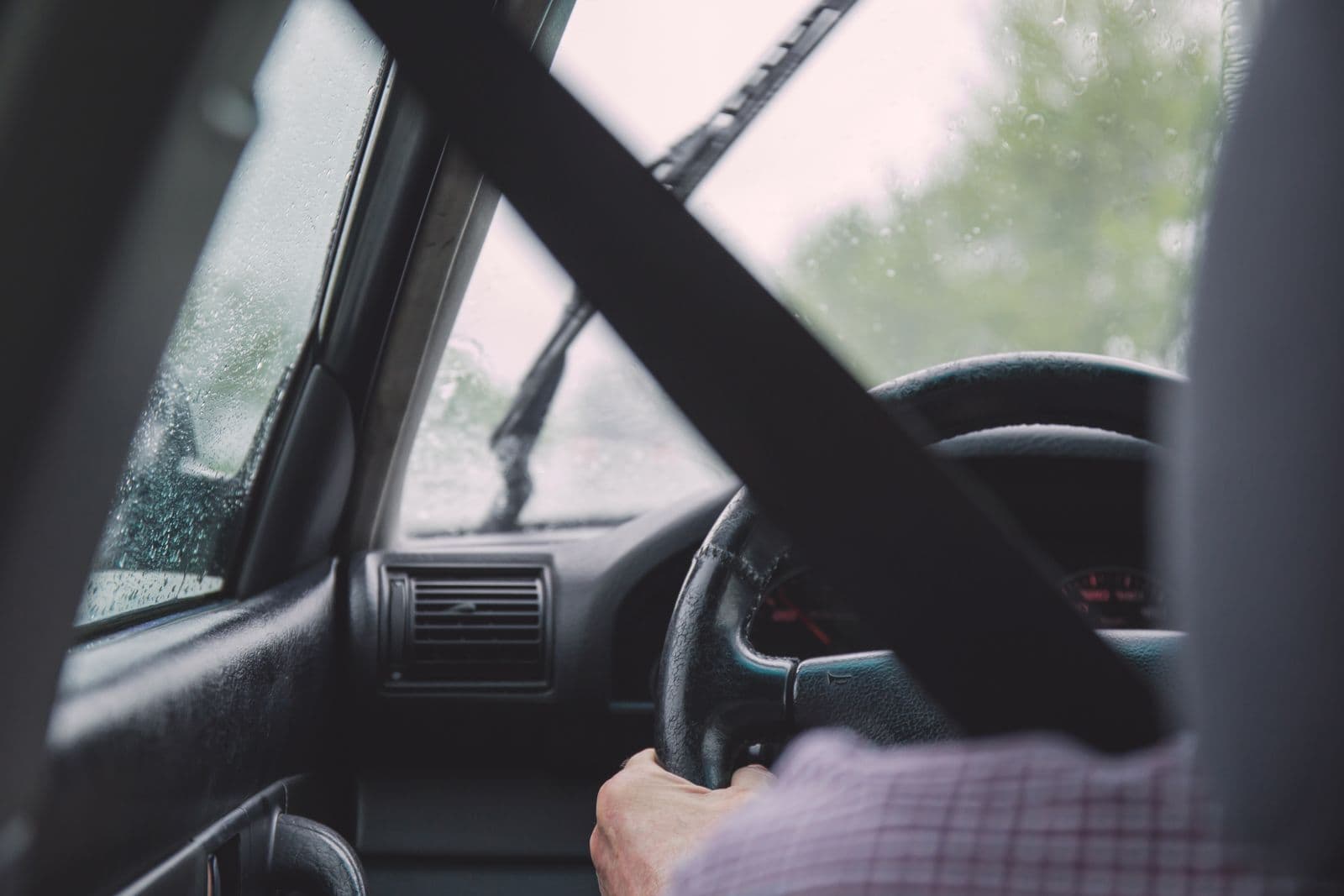 The view from a car's passenger seat of a seatbelted driver's hand on the steering wheel, with rain on the windscreen.