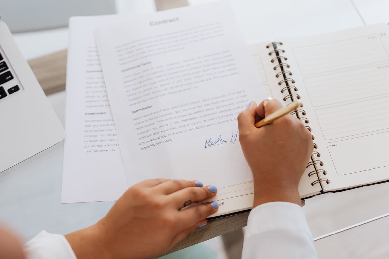 A hand signing the bottom of a printed contract on a desk beside an open notebook and a laptop.