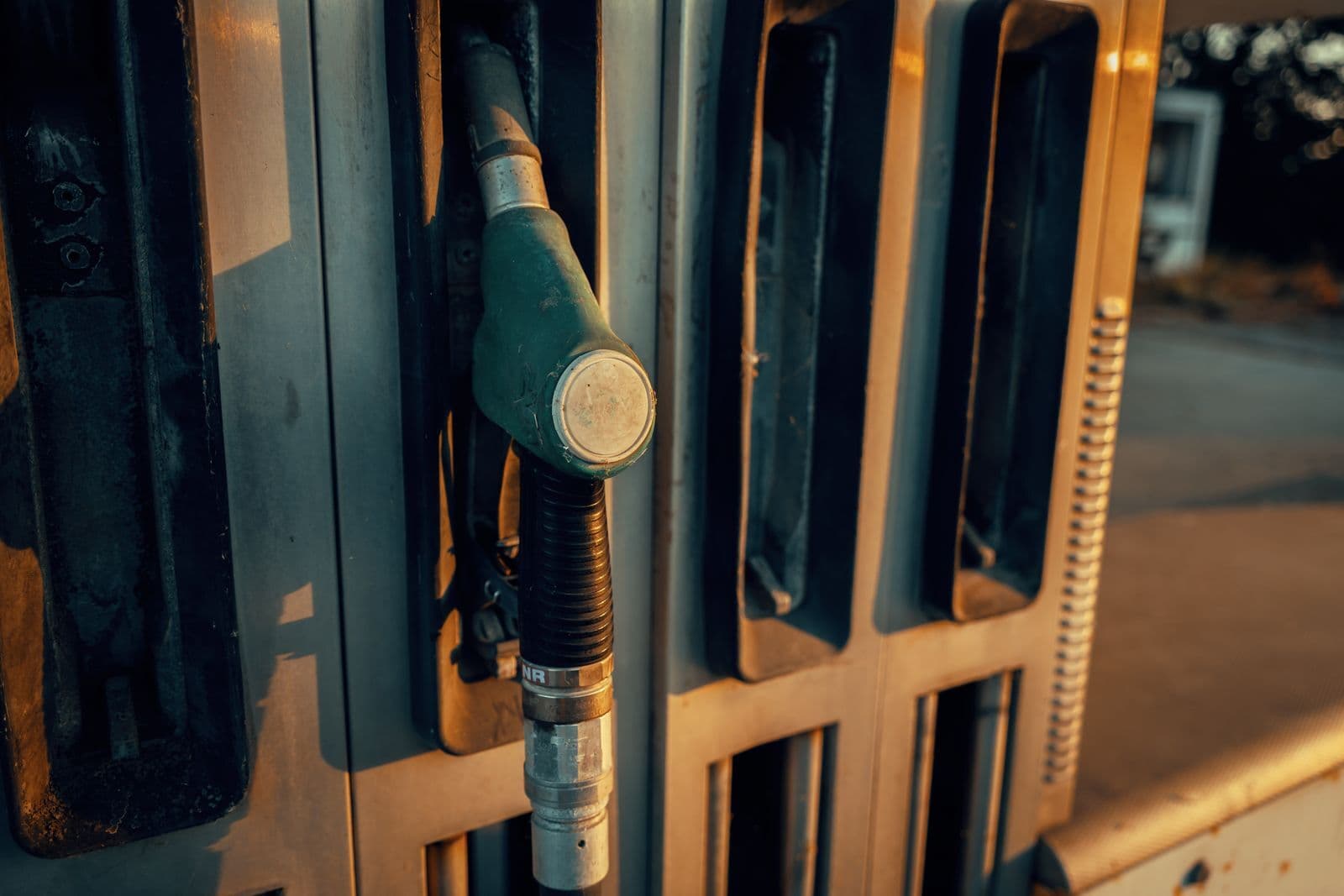 A weathered fuel nozzle resting in its holster on a service station pump, lit by low golden sunlight.