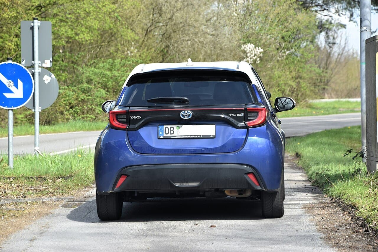 The rear of a blue Toyota Yaris Hybrid hatchback with German number plates, parked on a narrow country road.