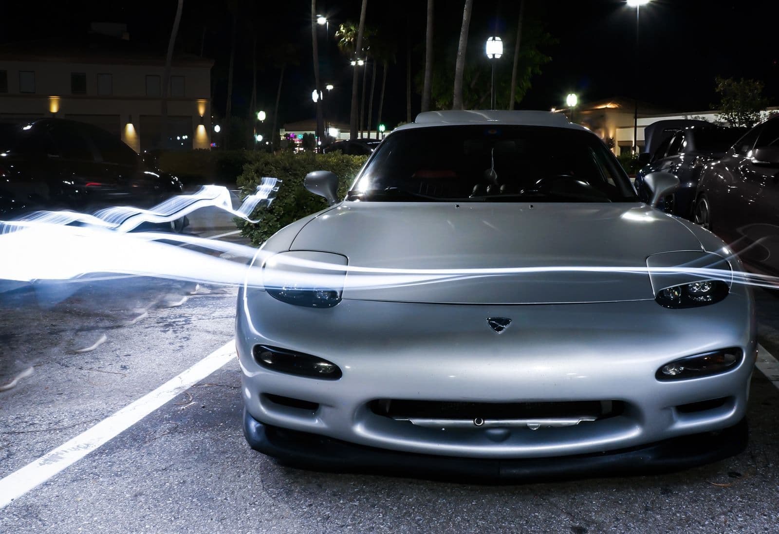 A silver Mazda RX-7 photographed head-on in a car park at night, with streaks of light trailing across the bonnet.