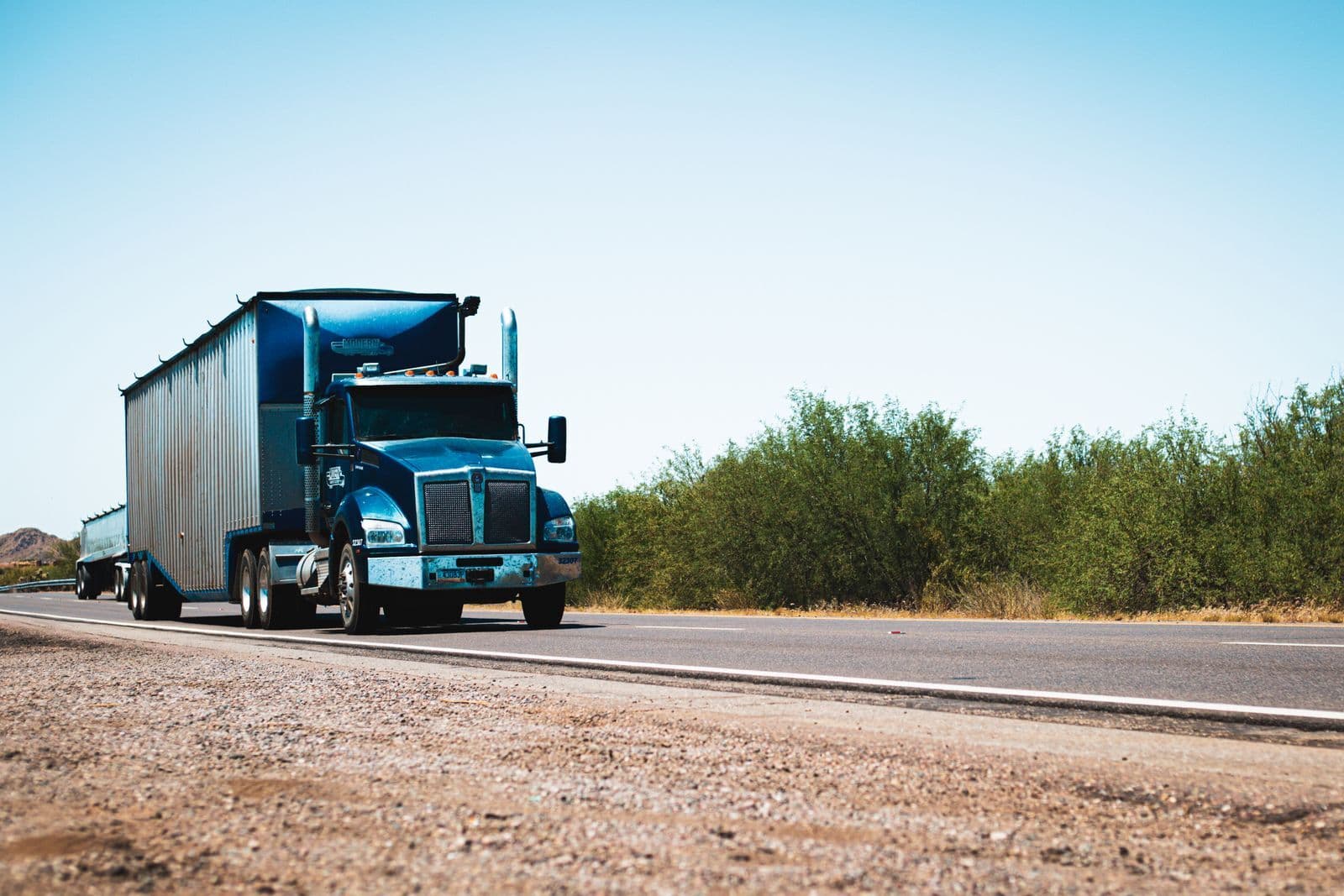 A blue prime mover towing two trailers along a highway through dry scrubland under a clear sky.