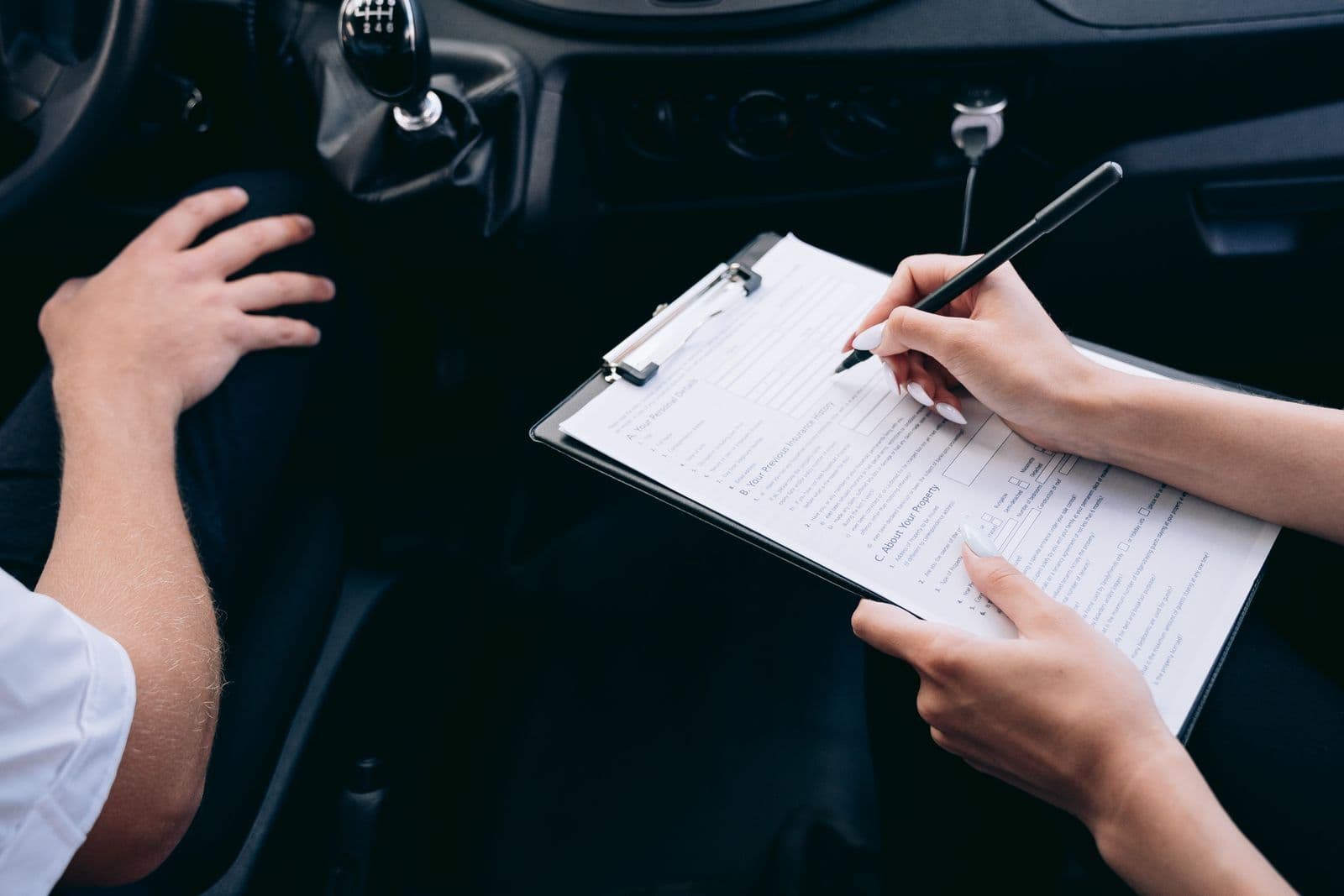 A hand filling in a form on a clipboard inside a car, beside the driver's hand resting on his knee.