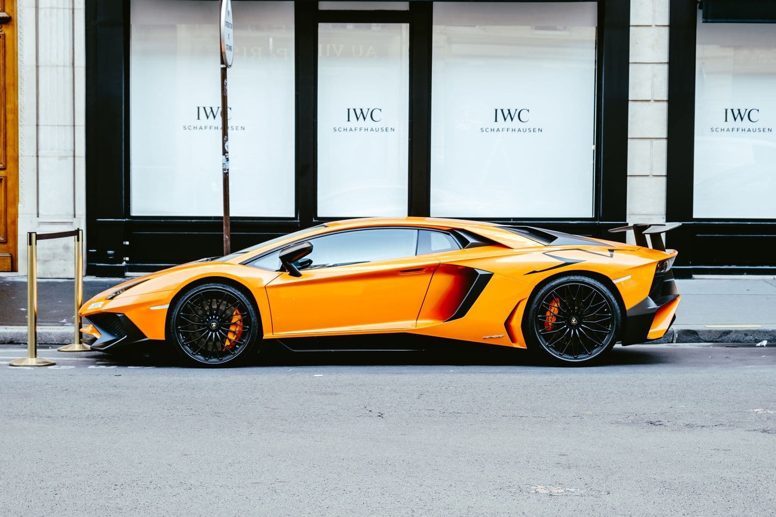 An orange mid-engined supercar with a fixed rear wing, parked in side profile on a city street.