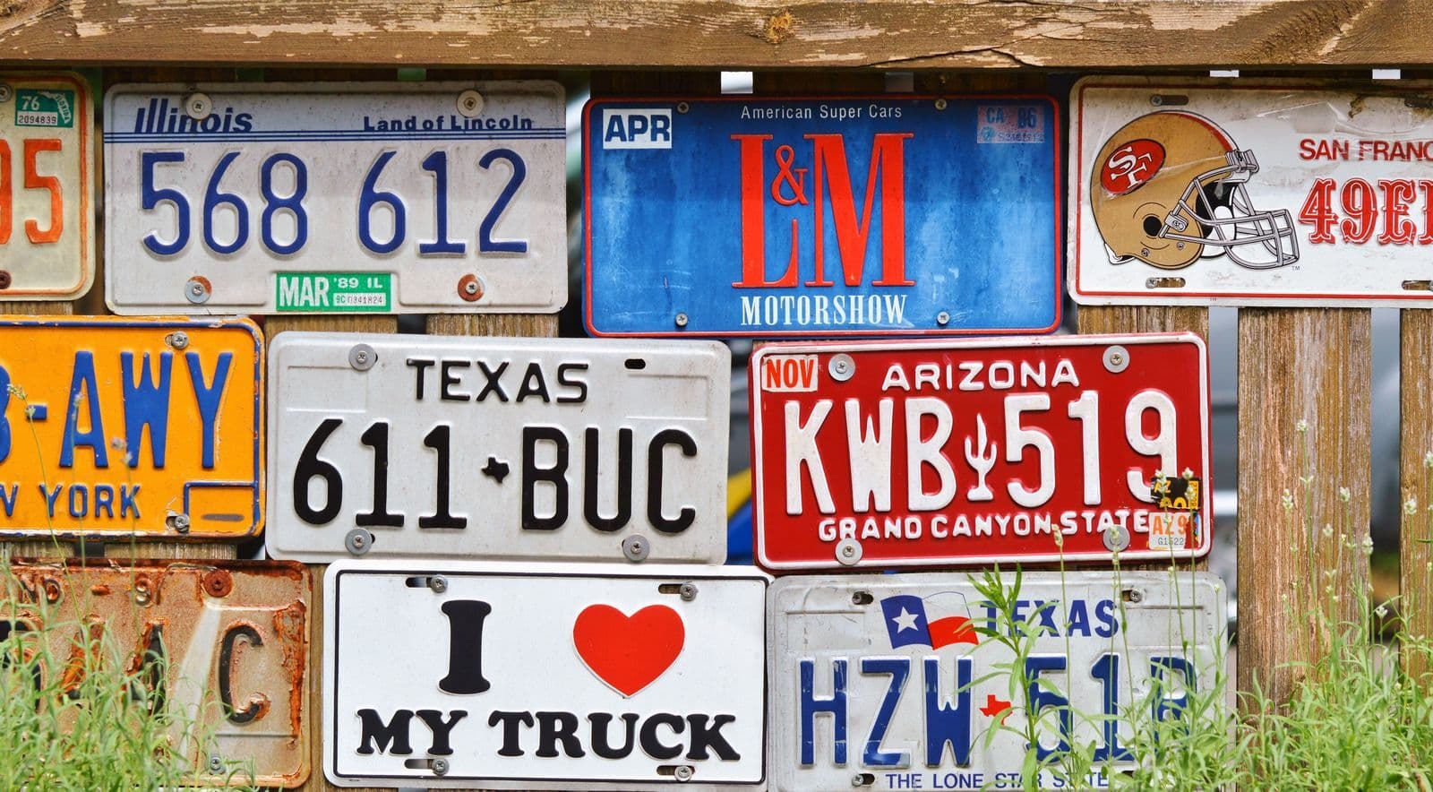 A weathered timber fence covered in dozens of old vehicle number plates in assorted colours and designs.