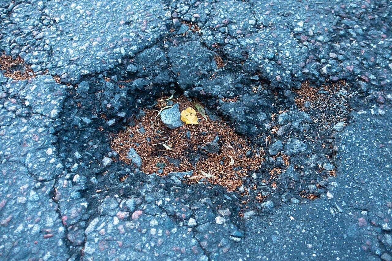 A large water-filled pothole in an asphalt road, its edges ringed by cracked and crumbling bitumen