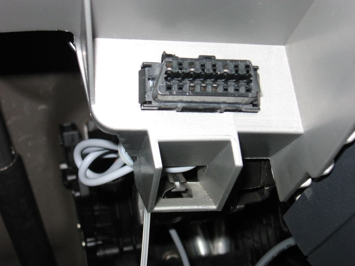 A vehicle OBD-II diagnostic socket under a dashboard, with its trapezoidal sixteen-pin connector