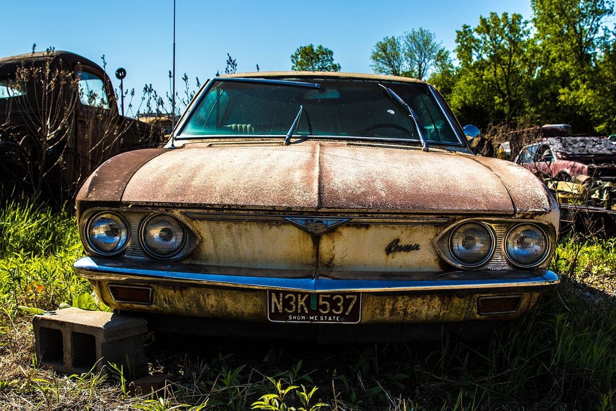 A corroded steel wheel arch and front fender on a car in a wrecking yard, with paint flaked away and the metal perforated along the arch lip