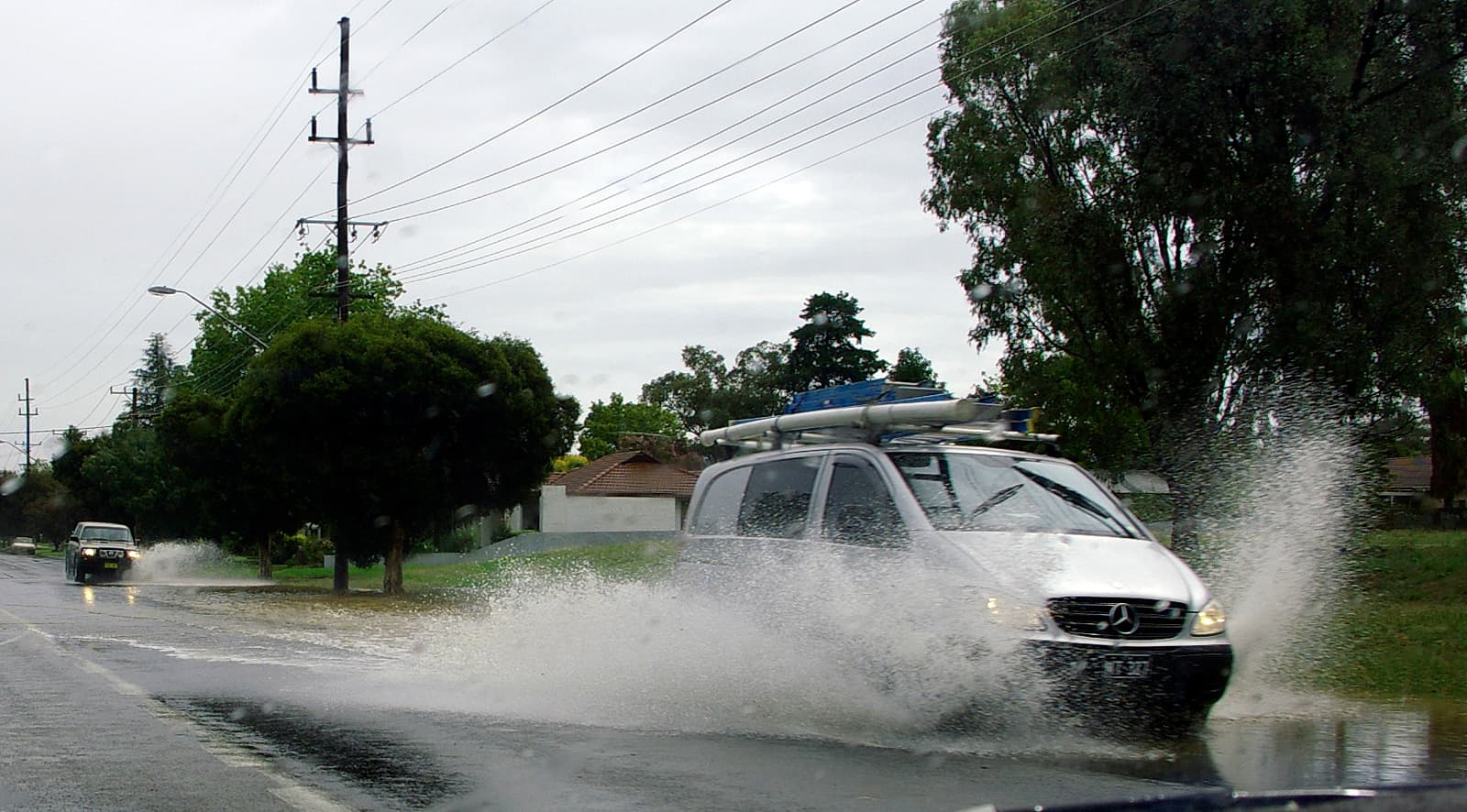 A white van throws up a wall of spray as it drives through flash-flood water across a suburban road, with a second car behind it.