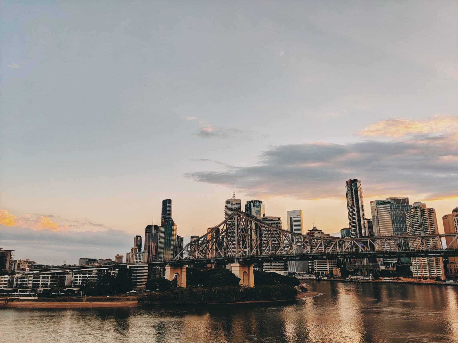 A steel truss bridge crossing a river in front of a city skyline at sunset.