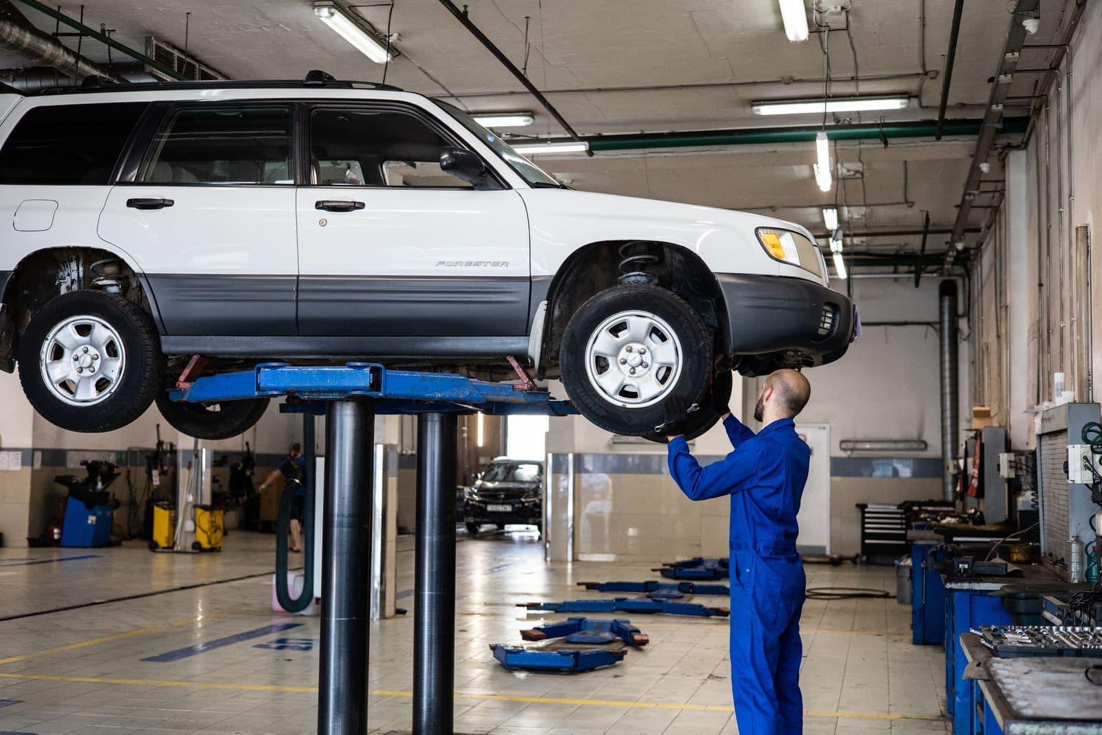 A white SUV raised on a workshop hoist while a mechanic in blue overalls inspects its front wheel.