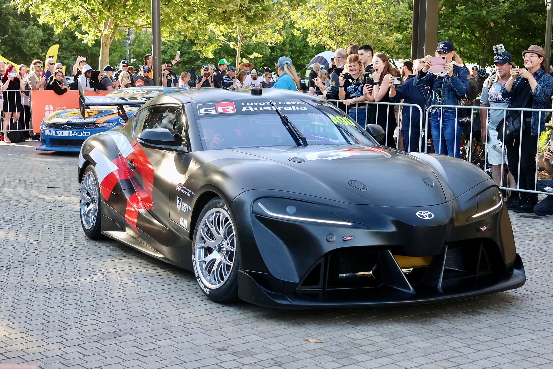 A black Toyota GR Supra Supercar race car on display in a city square with spectators behind barriers.