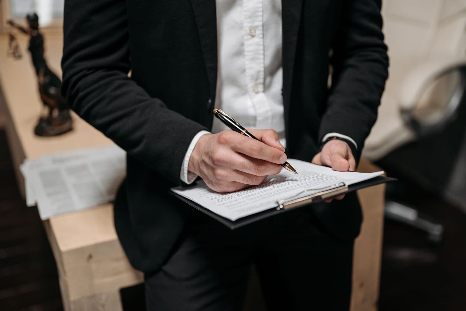 A man in a dark suit writes with a pen on papers held on a clipboard, with a white car beside him.
