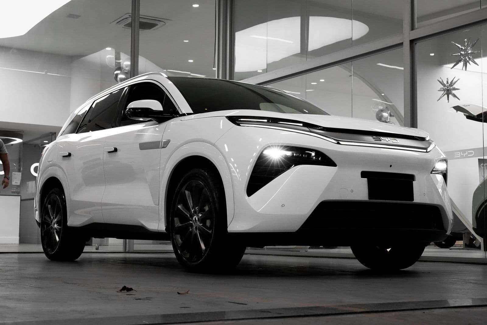 A white electric SUV with its headlights on, parked on the polished floor of a glass-walled showroom.