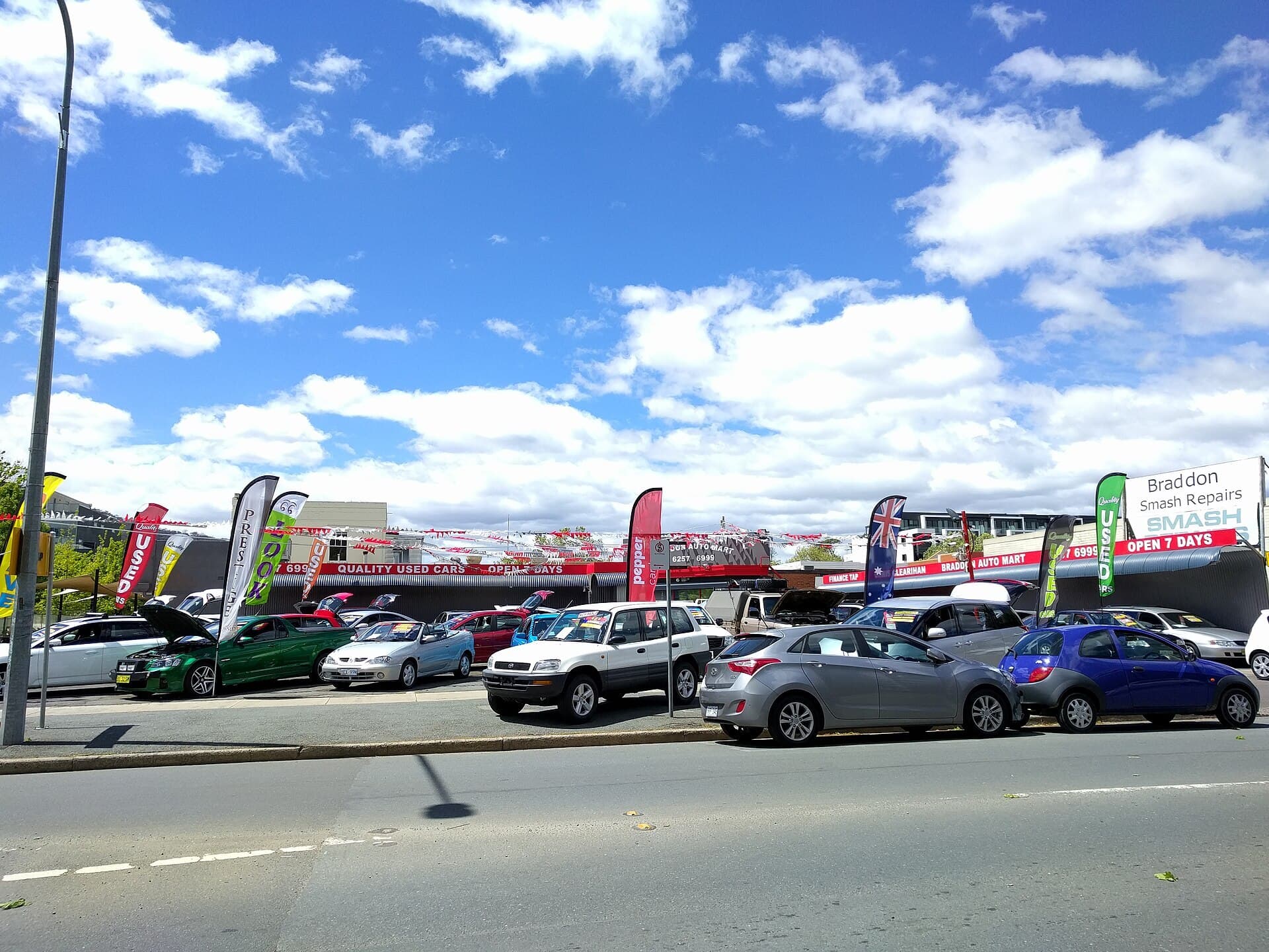 A second-hand car yard with rows of parked cars, bunting and flags reading Used under a blue sky with white clouds.