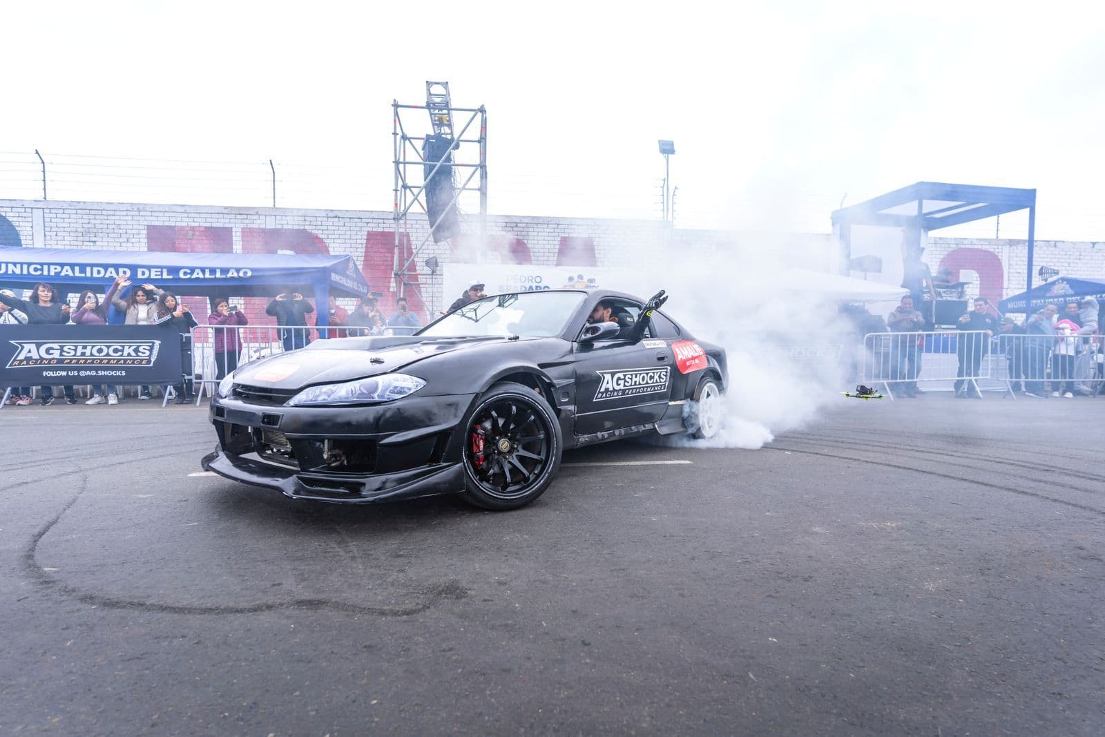 A black modified coupe spinning its rear wheels in a cloud of tyre smoke in front of spectators behind a barrier.