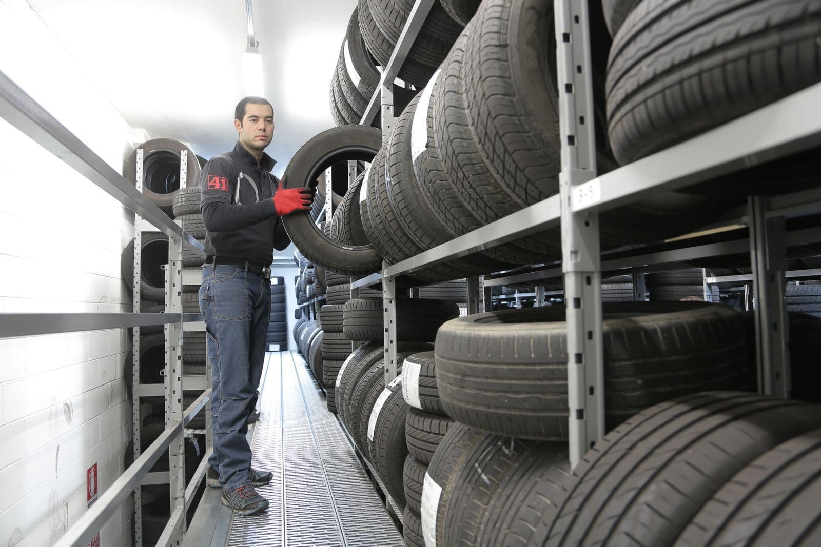 A worker lifting a tyre onto tall metal racks stacked with new tyres in a storage room.