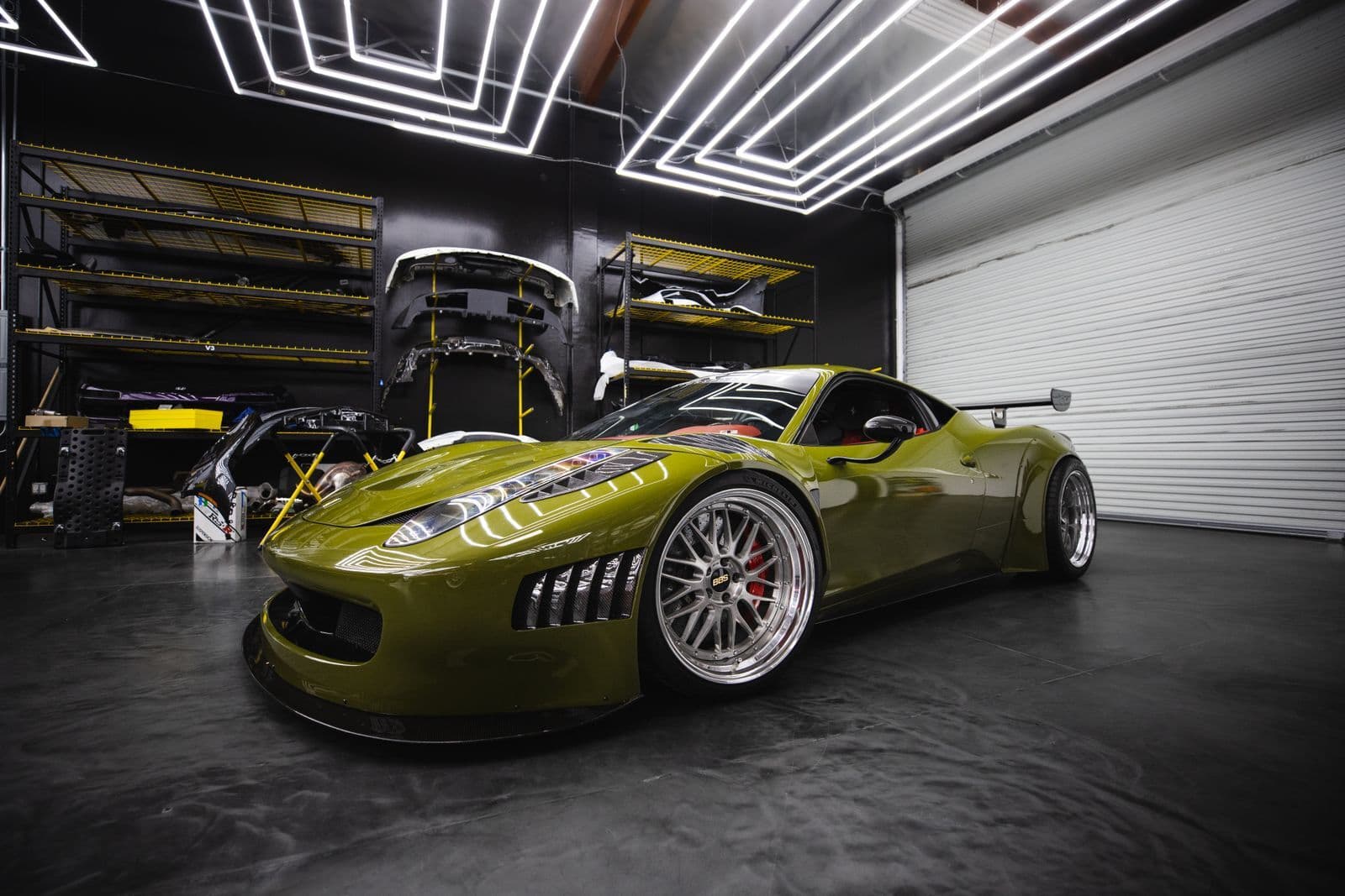 A green wide-bodied mid-engined sports car with a rear wing parked in a workshop, with spare body panels on racking behind it.