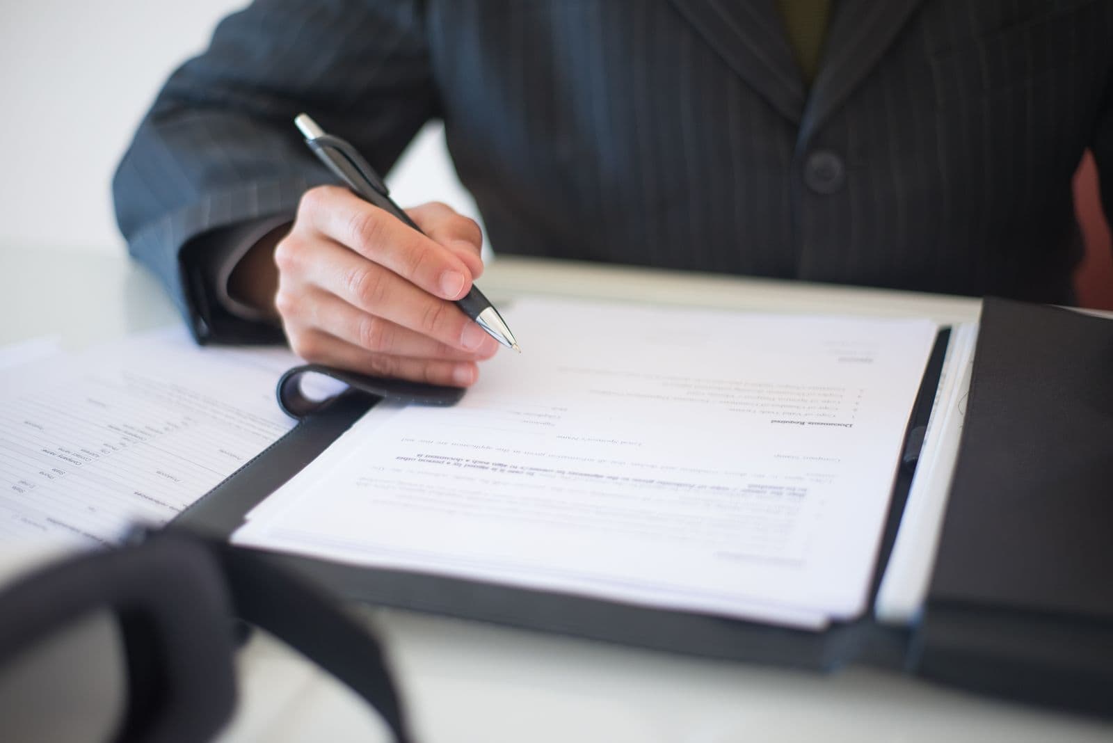 A man in a pinstripe suit holds a pen over a printed contract in a black folder on a desk.