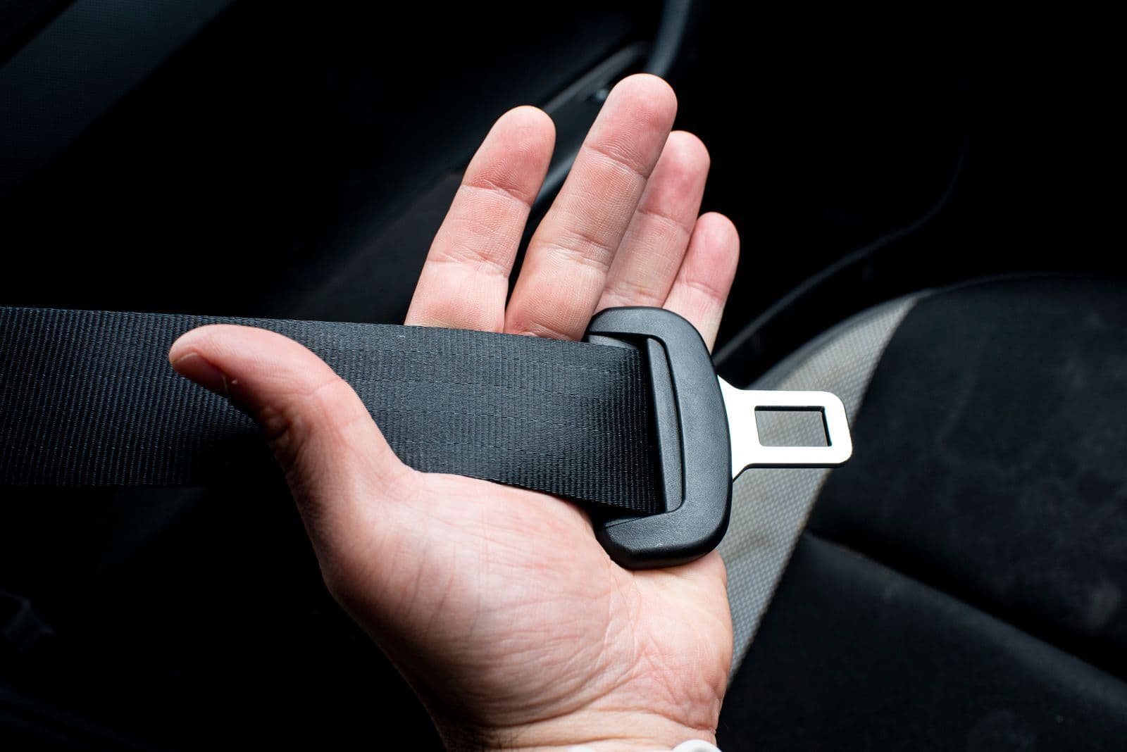 A hand holding a black car seat belt and its metal buckle tongue beside a fabric seat.