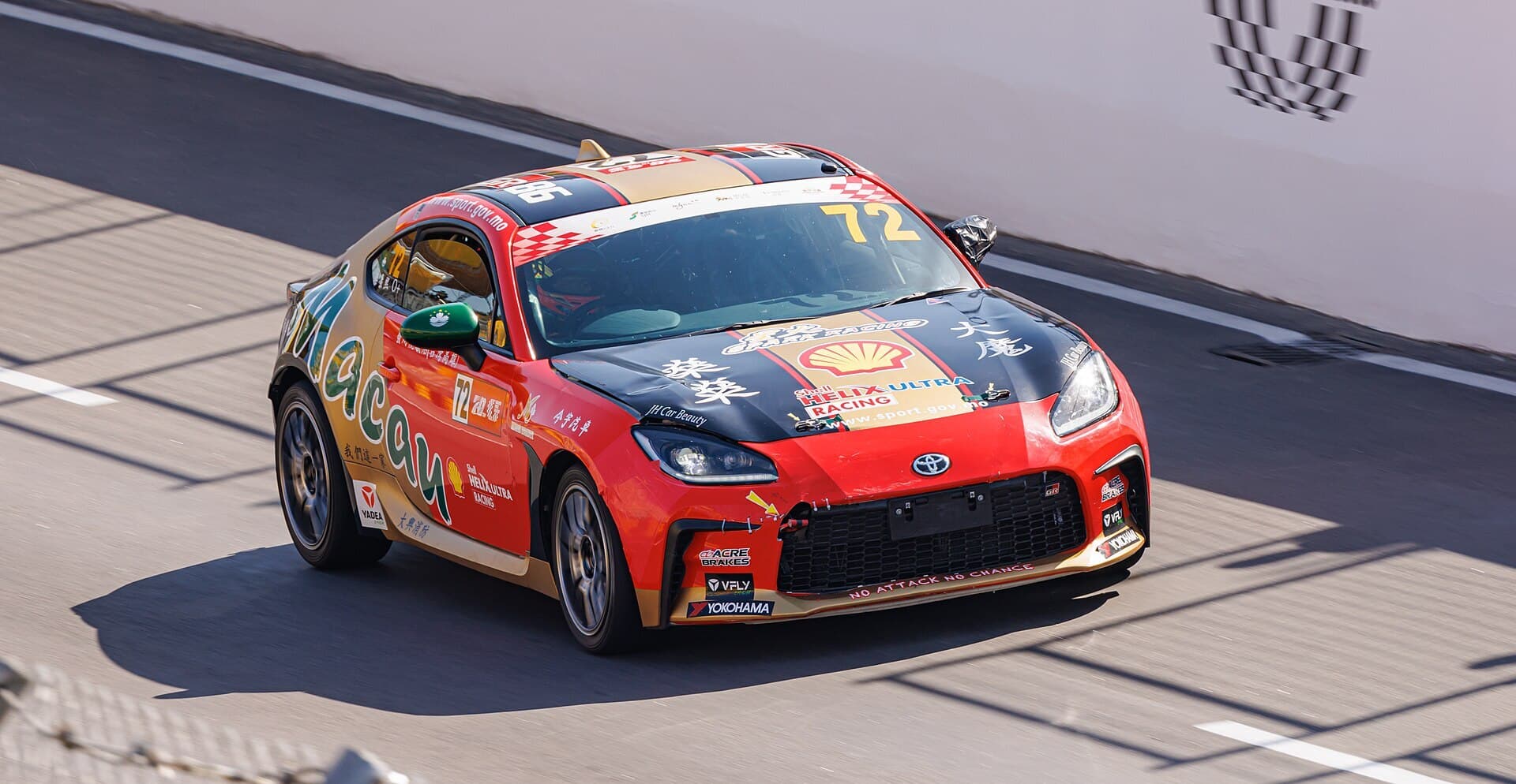A red and black Toyota GR86 race car cornering beside a concrete wall on a street circuit.